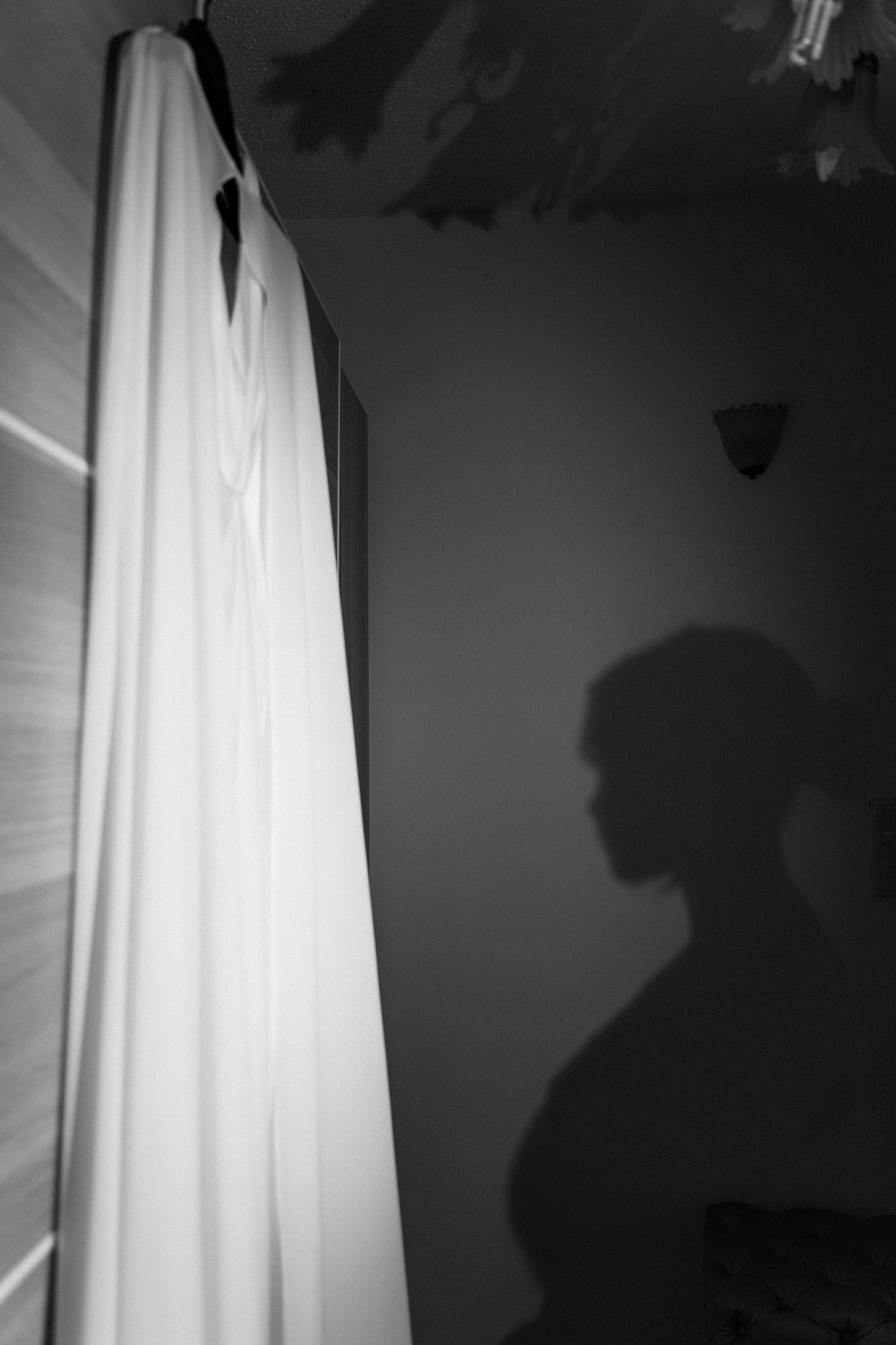 Black and white photo of white wedding dress hanging beside silhouette shadow on wall.