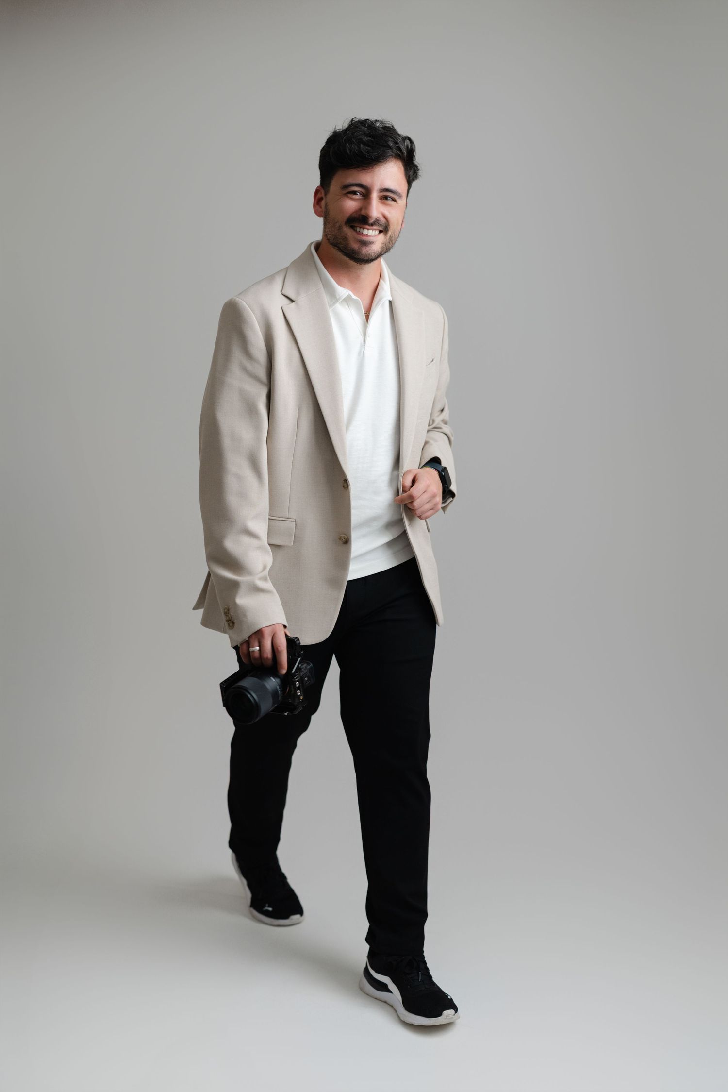 Smiling man in beige blazer and black pants poses confidently holding a camera in studio.