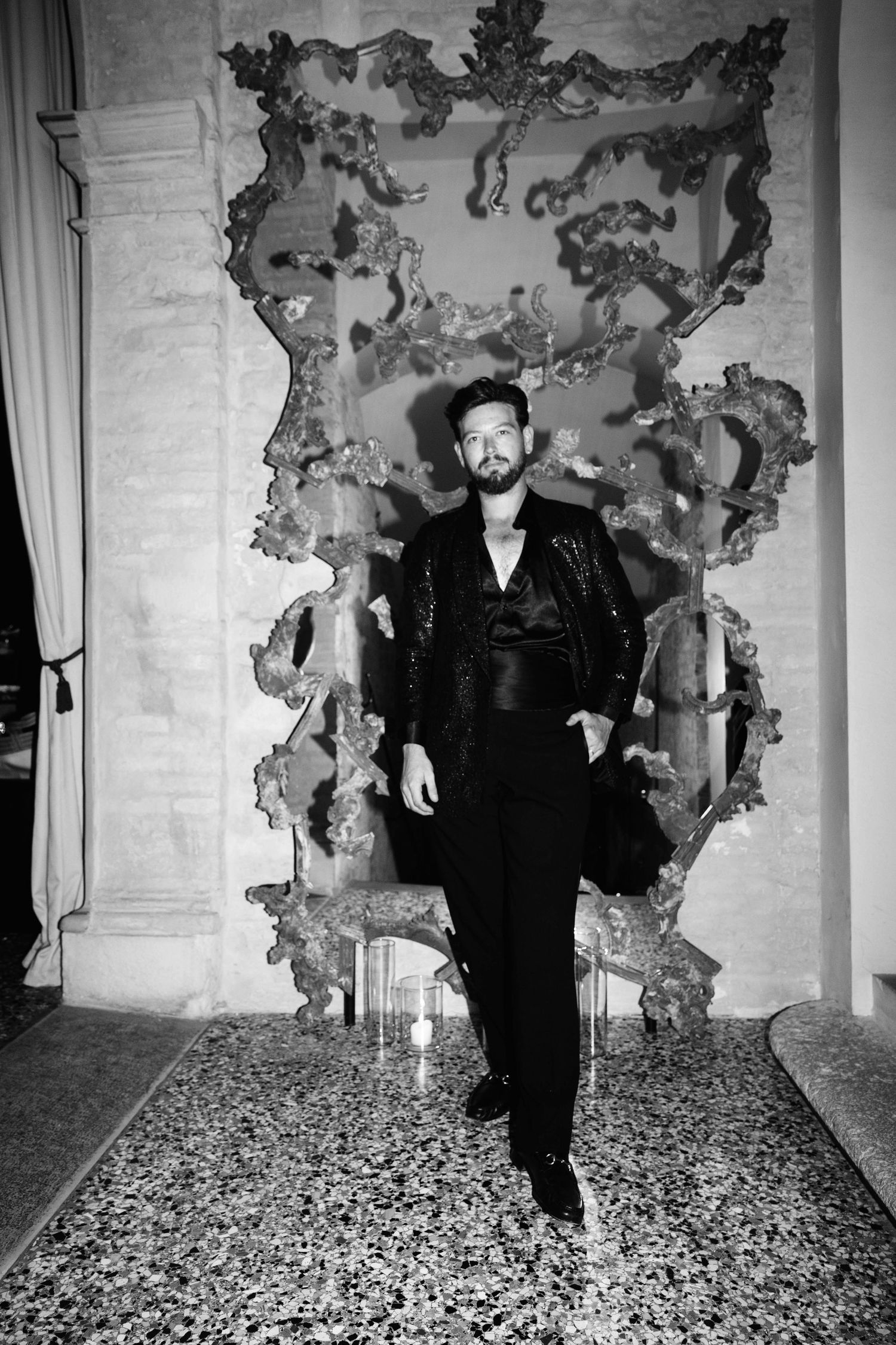 Stylish man in black suit posing before ornate baroque mirror in black and white photo.