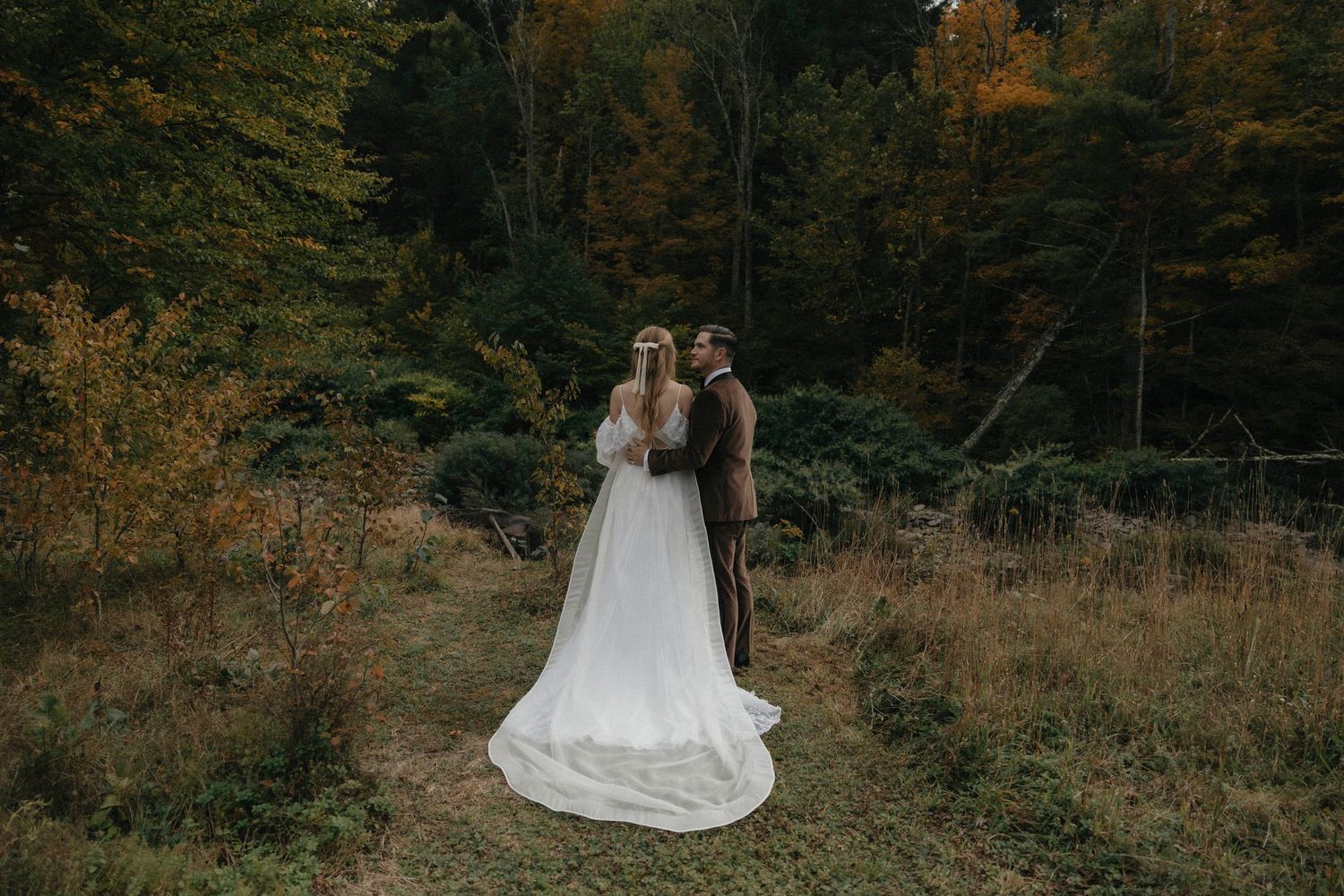 Bride and groom walking hand-in-hand through colorful autumn forest in wedding attire.