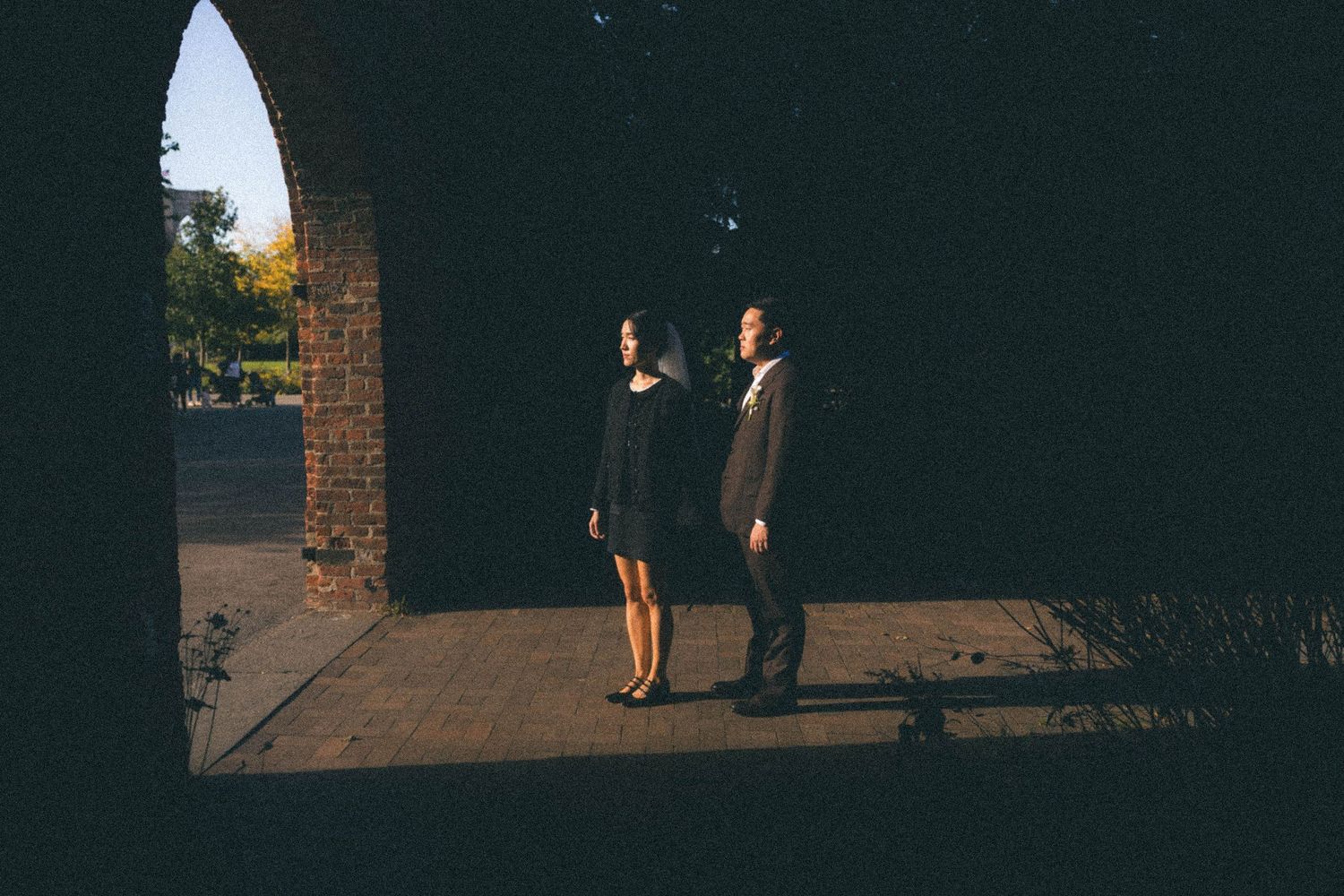 Bride and groom stand in golden light beneath brick archway in artistic wedding portrait.