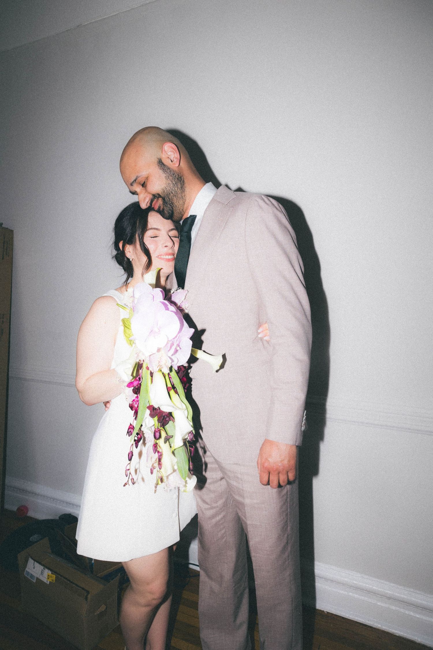 Groom in tan suit tenderly kisses bride's forehead as she holds floral bouquet indoors.