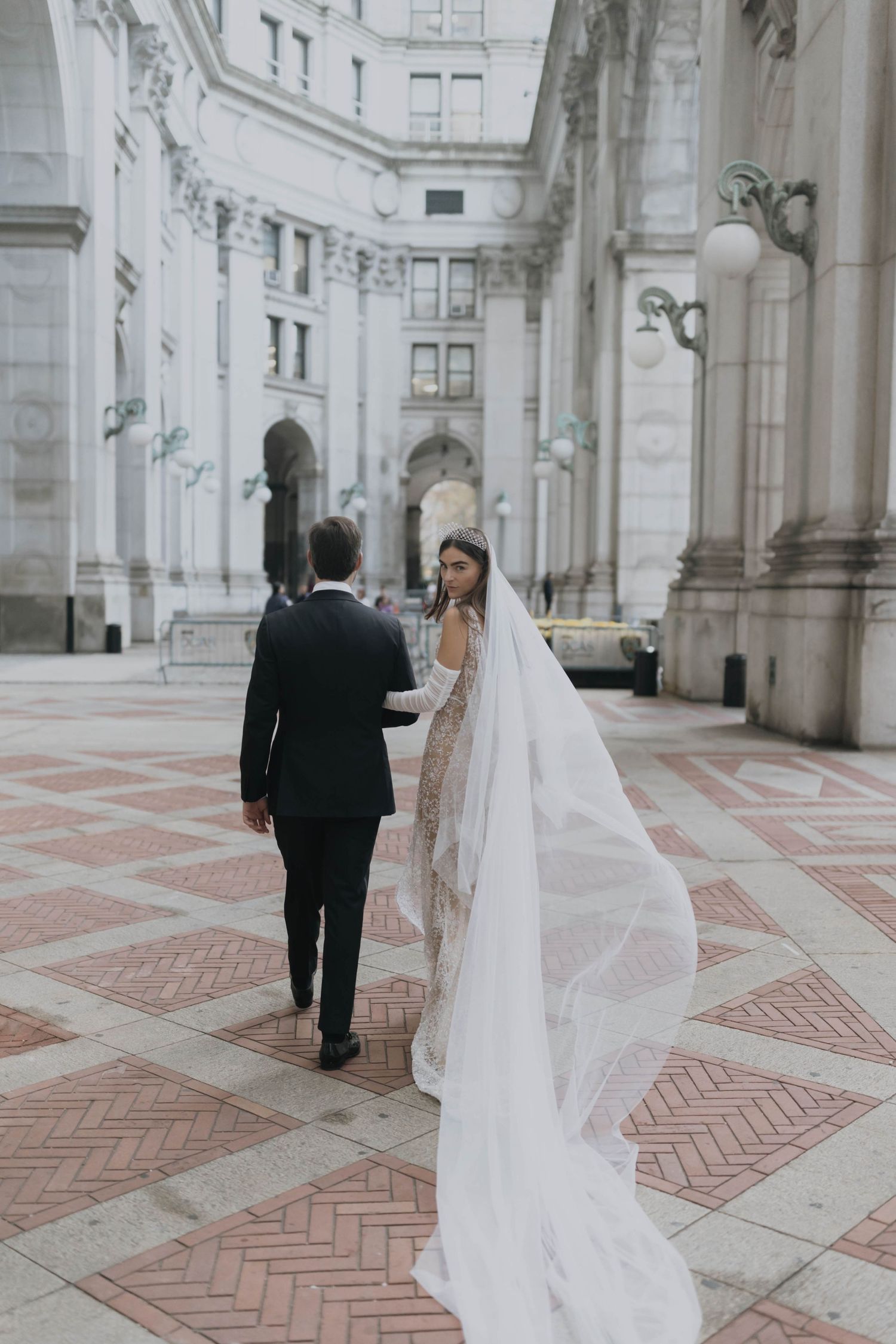 Bride and groom walk through elegant neoclassical courtyard, her long veil trailing across brick pavers.