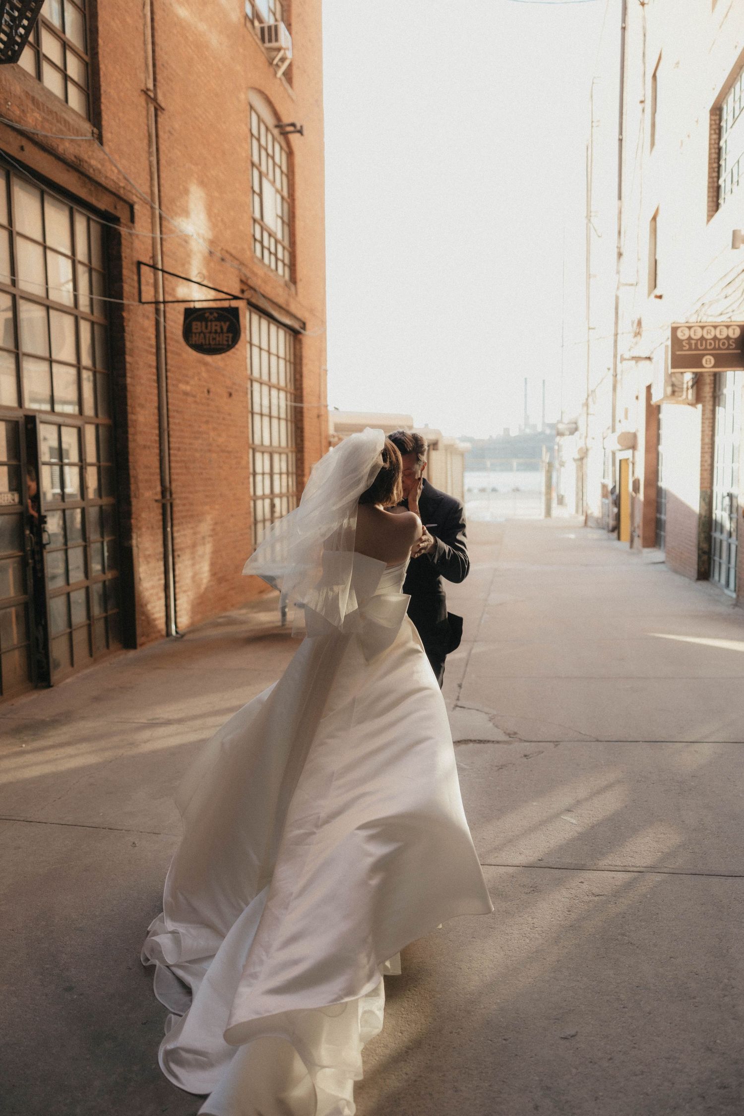 Bride and groom walking together along sunlit urban alley beside brick building on wedding day.