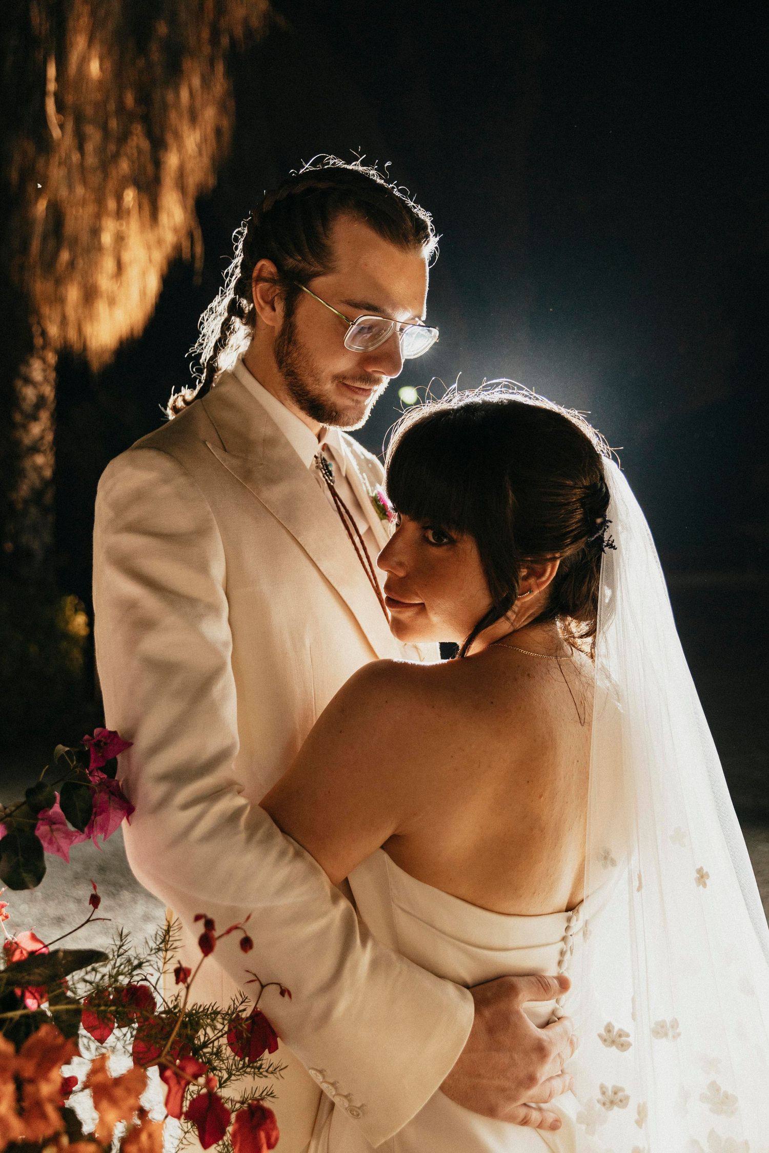 Groom in cream suit tenderly embraces bride in strapless gown under a dramatic spotlight at night.