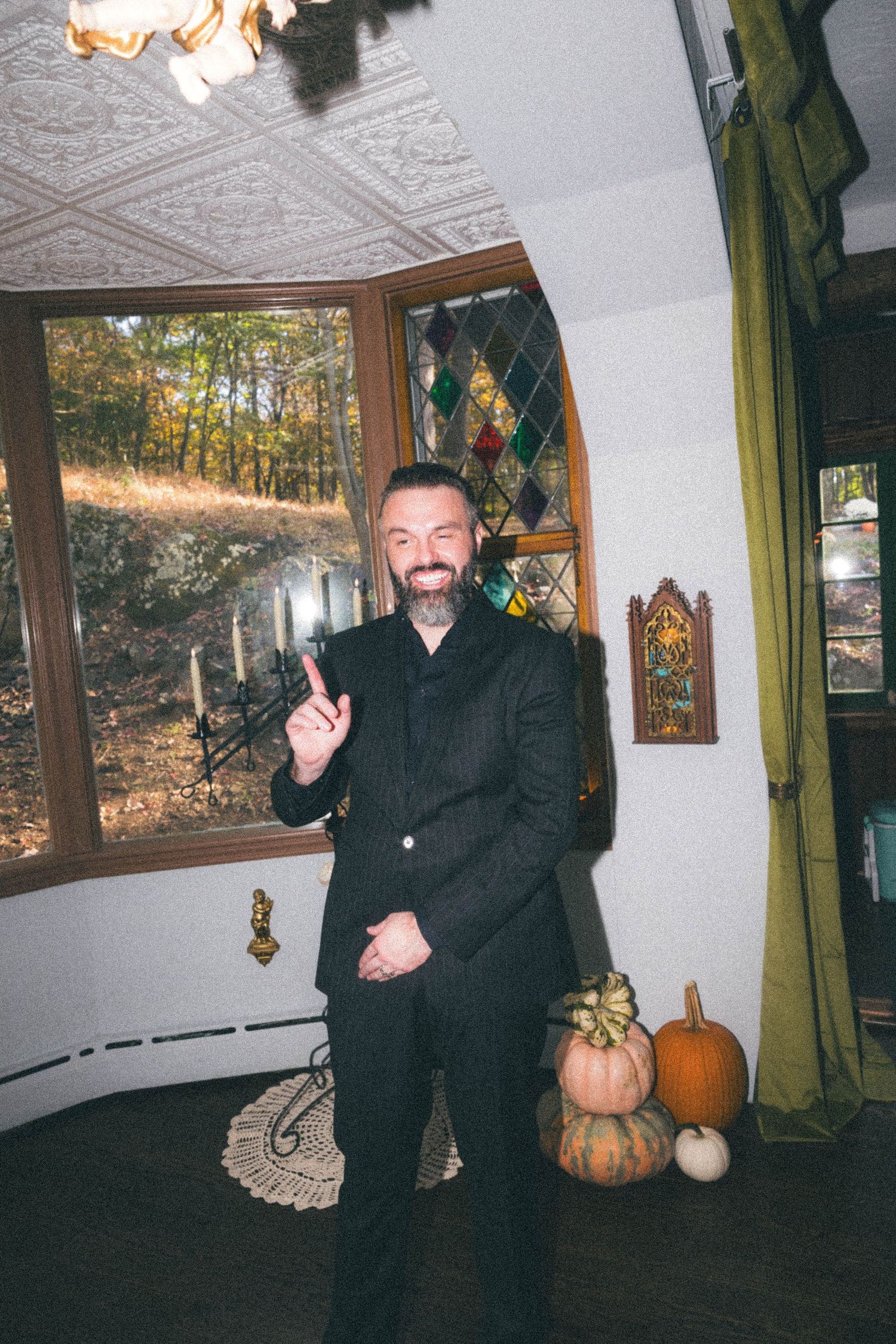 Smiling bearded man in black suit giving thumbs up near stained glass window with Halloween pumpkins.