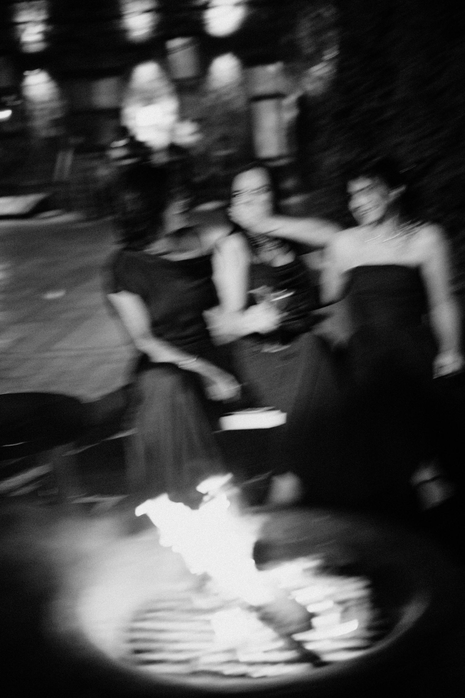 Blurred black and white photo of women in formal gowns dancing around outdoor fire pit.