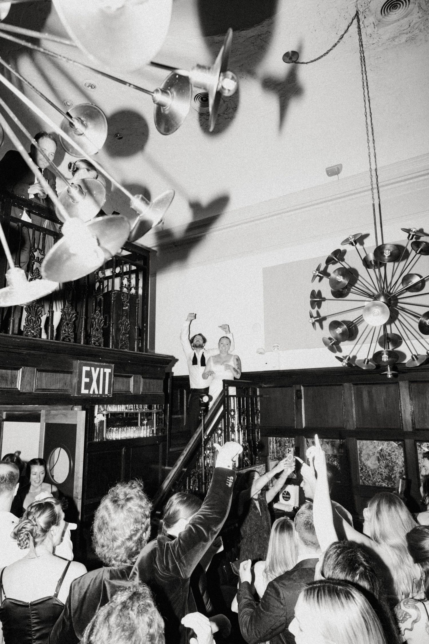 Black and white photo of lively wedding reception crowd cheering in elegant venue.