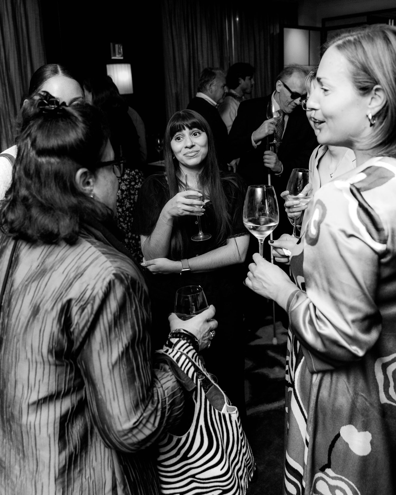 Black and white photo of women chatting and holding wine glasses at an indoor evening reception, one carrying a zebra-print bag.
