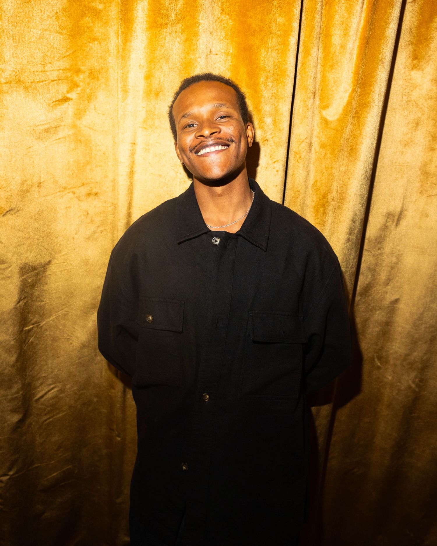 Smiling man in a black button-up shirt standing in front of a shimmering gold velvet curtain.