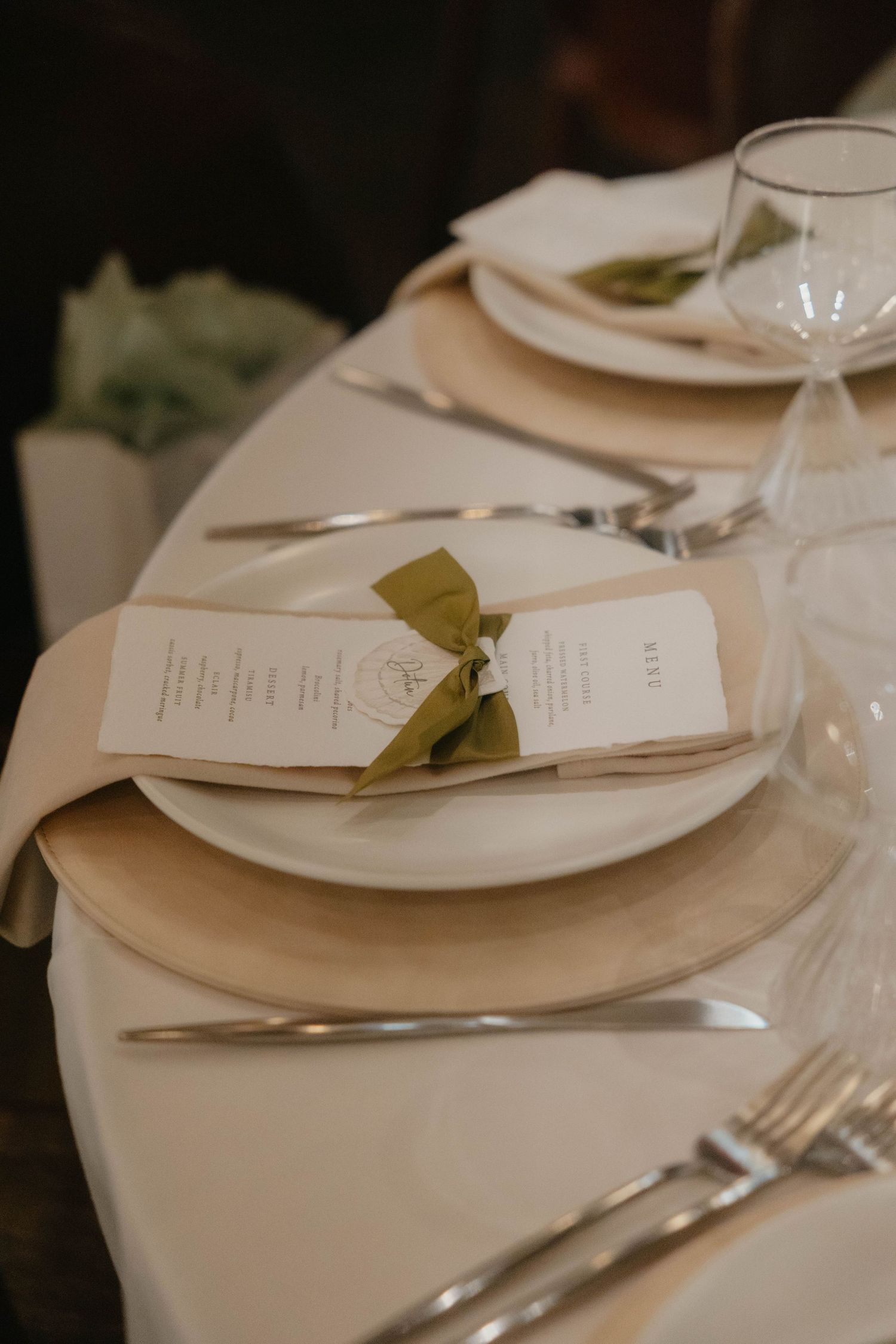 Elegant wedding table setting with a printed menu tied with an olive green ribbon on a white plate, gold charger, silverware and glassware.