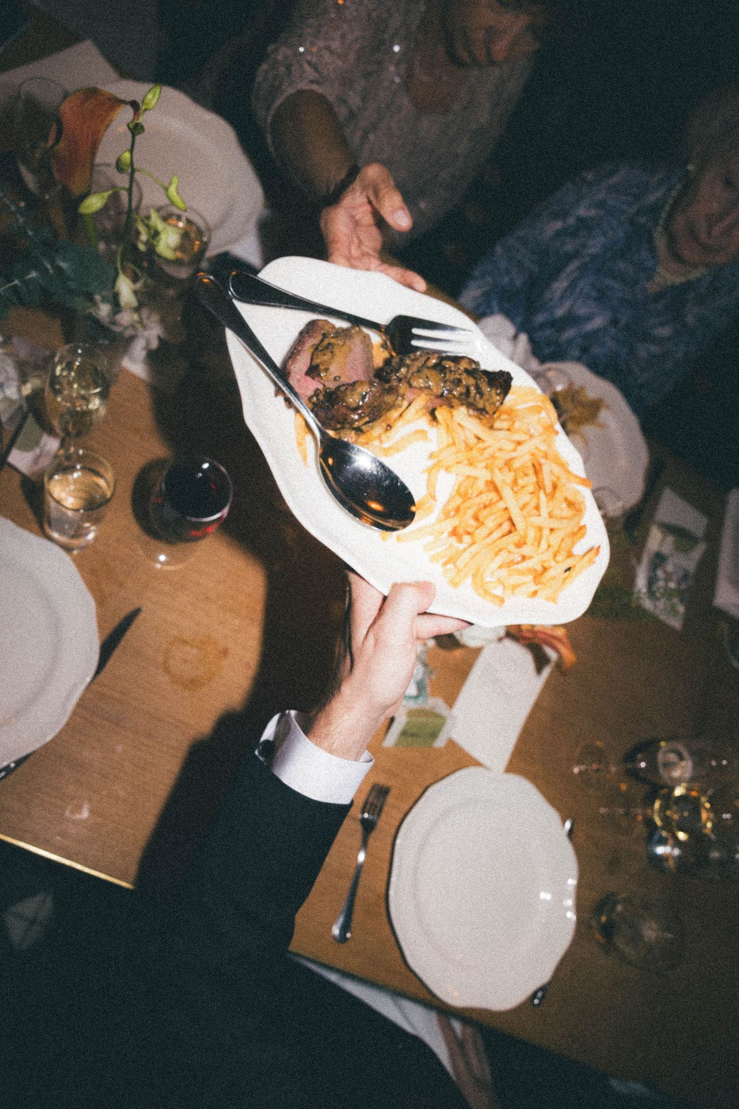 Wedding guest holds a platter of steak and french fries over a candlelit reception dinner table.