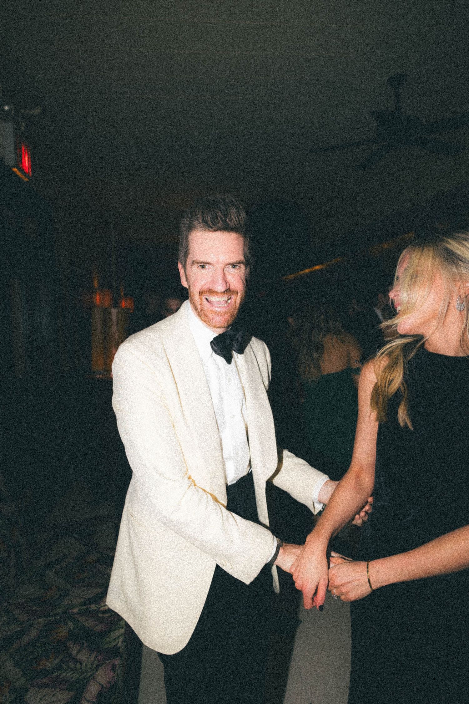 Groom in white tuxedo jacket and black bow tie smiling while dancing with partner at wedding reception.