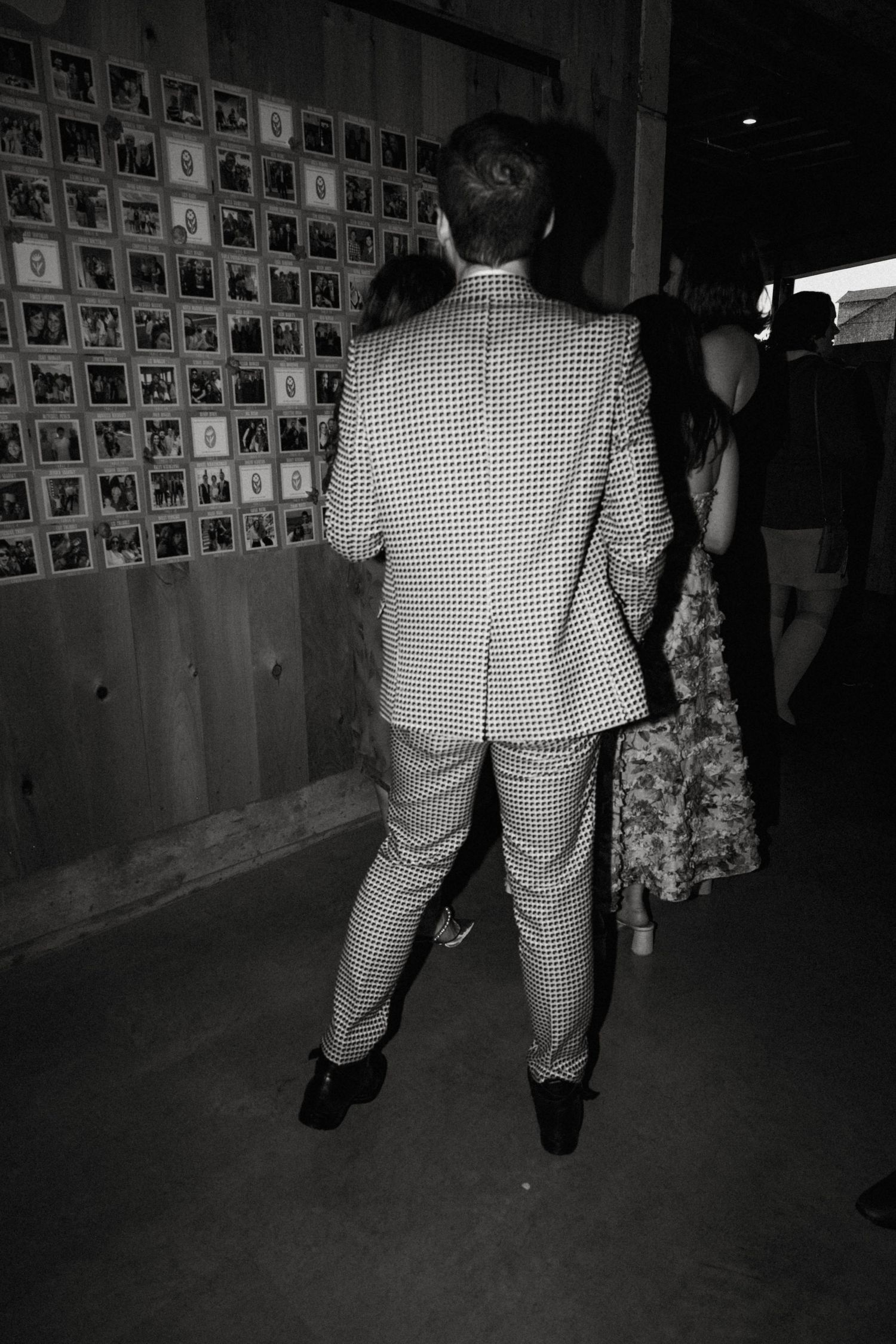 Black and white photo of a man in checkered suit viewing a photo display wall at a wedding reception.