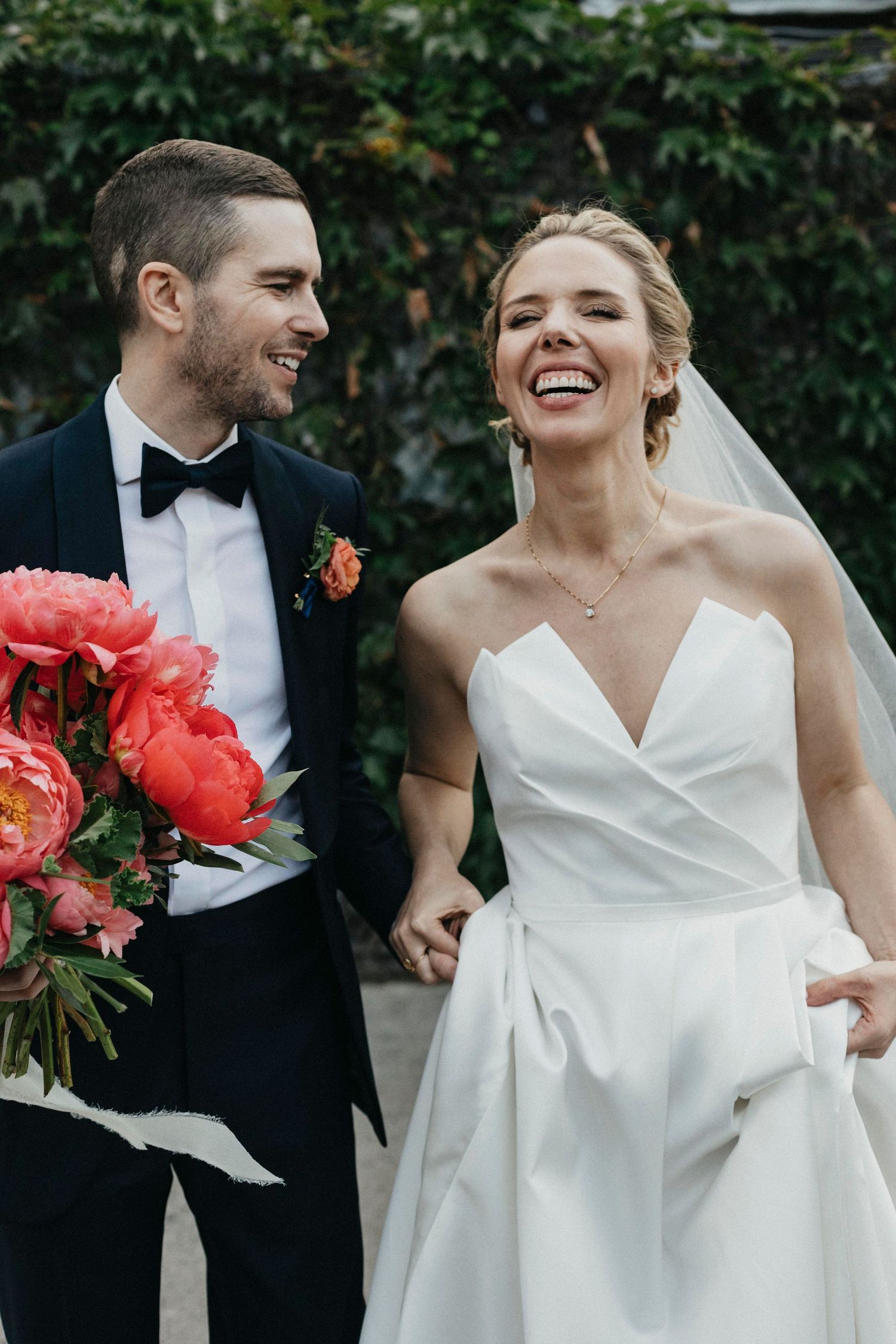 Smiling bride and groom walking together, groom in black tuxedo holding vibrant coral peony bouquet.