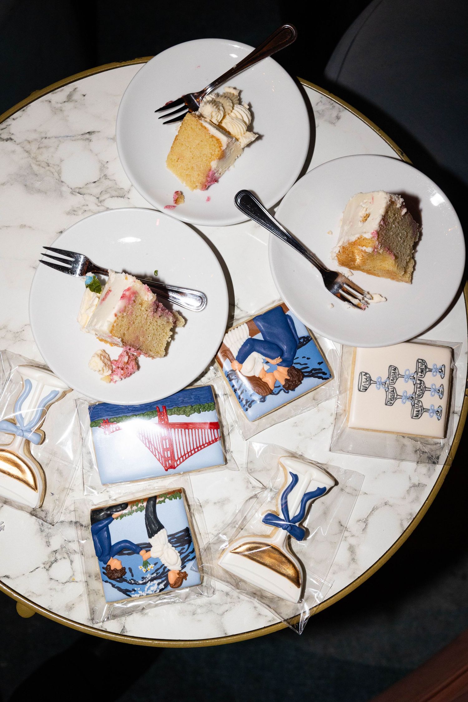 Overhead view of marble table with sliced wedding cake pieces on plates and decorated sugar cookies.