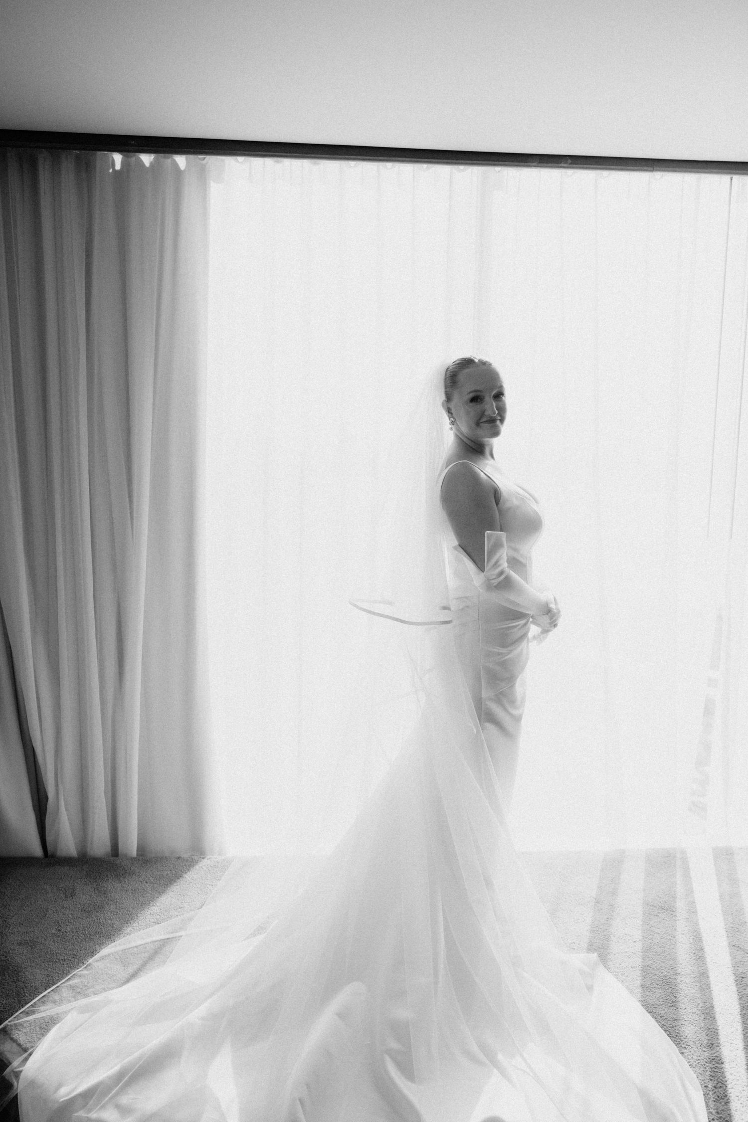 Black and white portrait of bride in fitted gown with veil standing before sheer curtains.