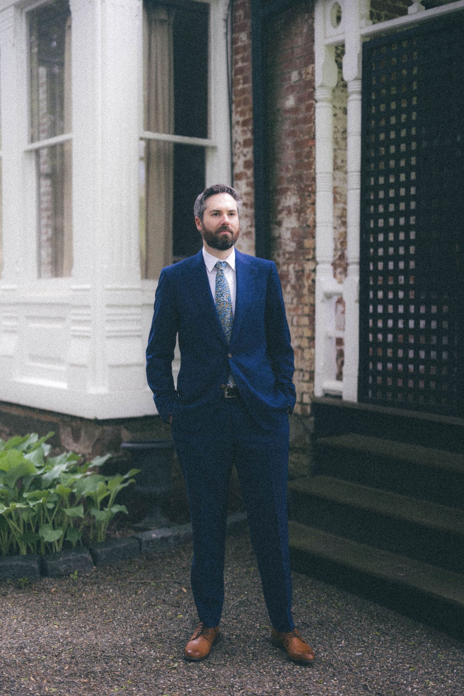 Bearded groom in navy blue suit and brown shoes standing confidently outside historic brick building.