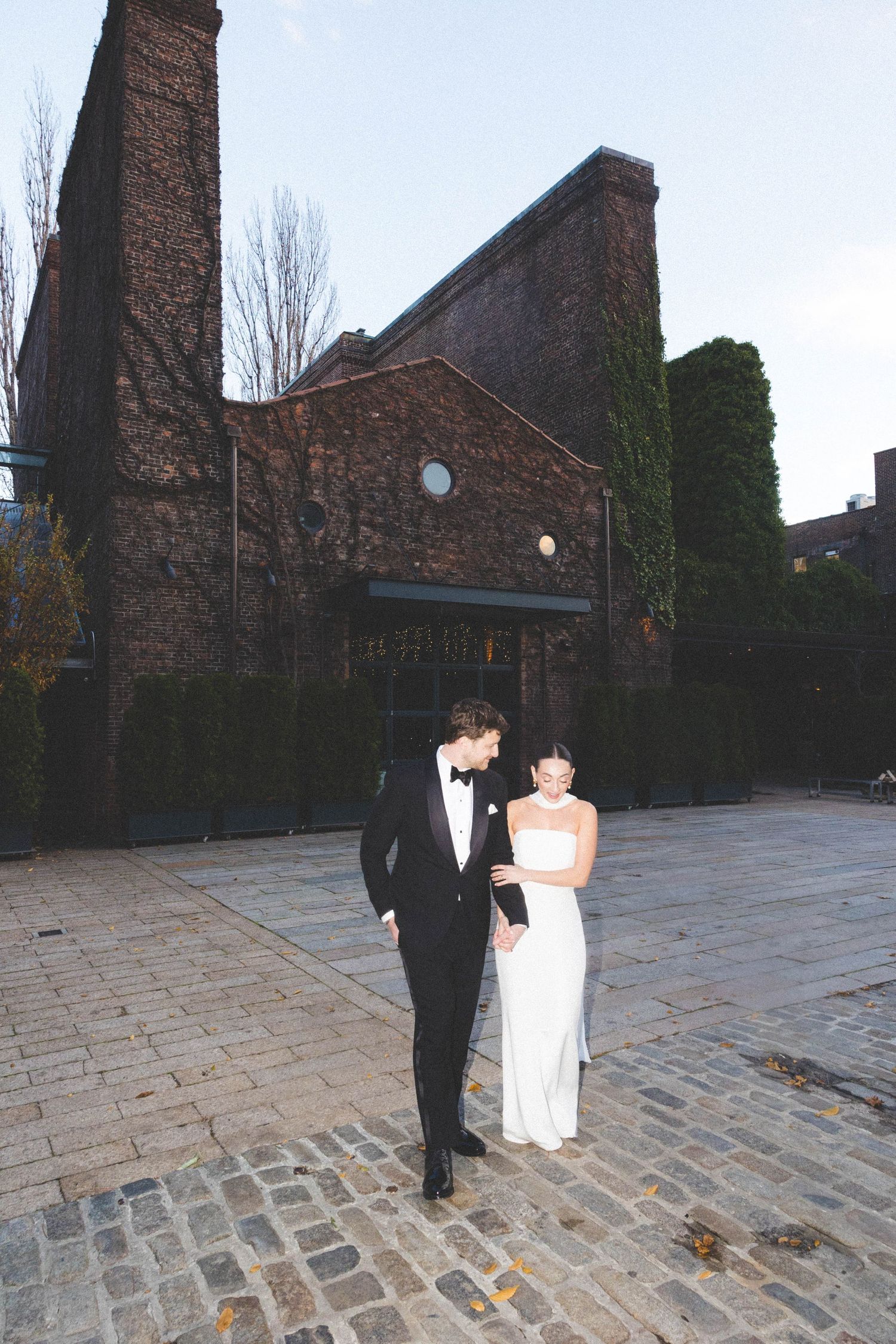 Bride and groom walking on cobblestone street outside historic brick building at dusk.