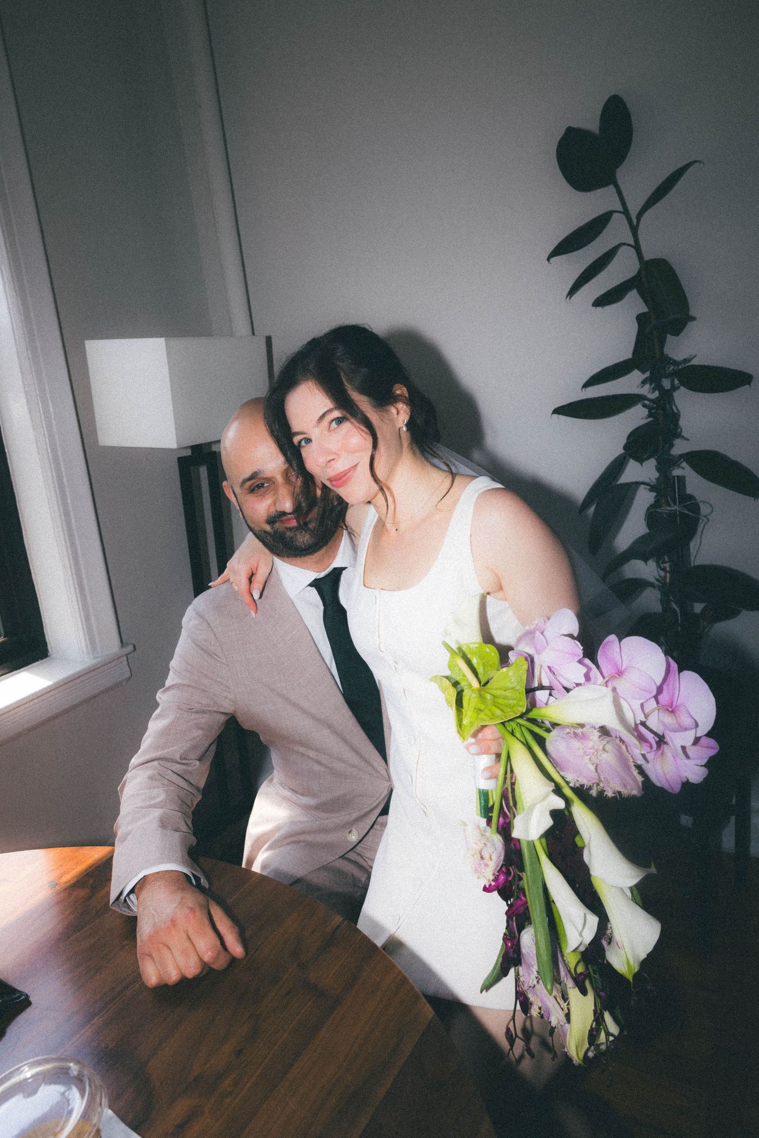 Bride in white dress with floral bouquet leans on seated groom at wooden table indoors.