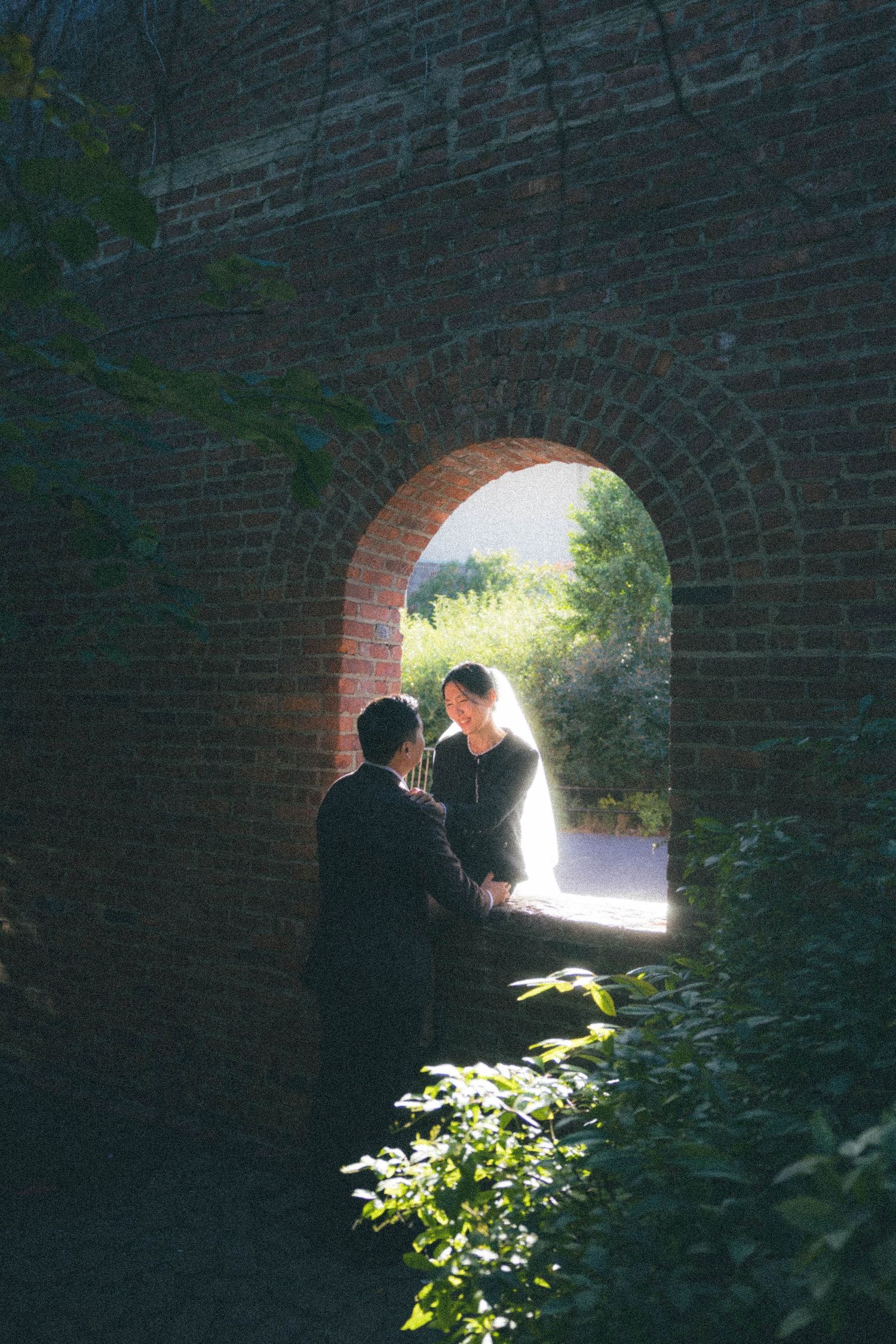 Bride and groom share tender moment framed by sunlit brick archway surrounded by greenery.