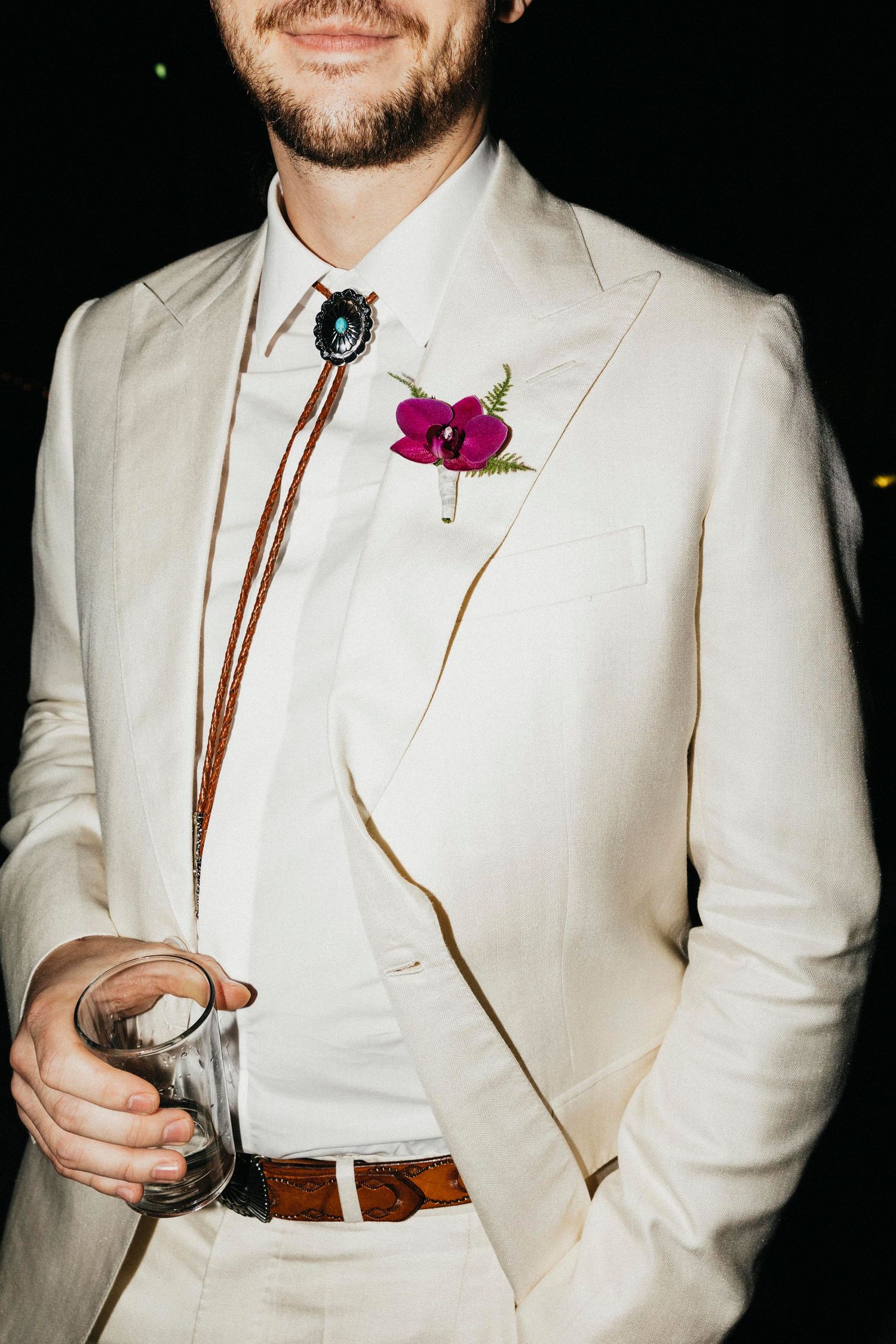 Groom in cream suit wearing a bolo tie and purple orchid boutonniere holds a glass at reception.