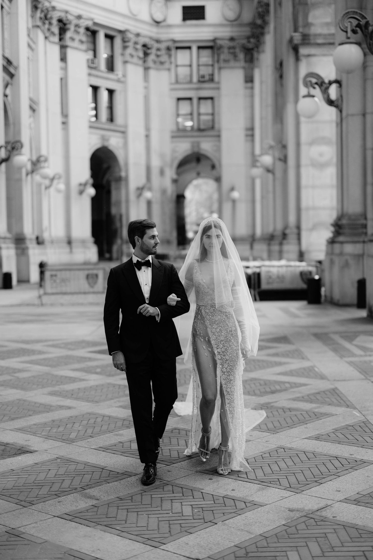 Black and white full-length photo of elegant couple walking through grand neoclassical building interior.