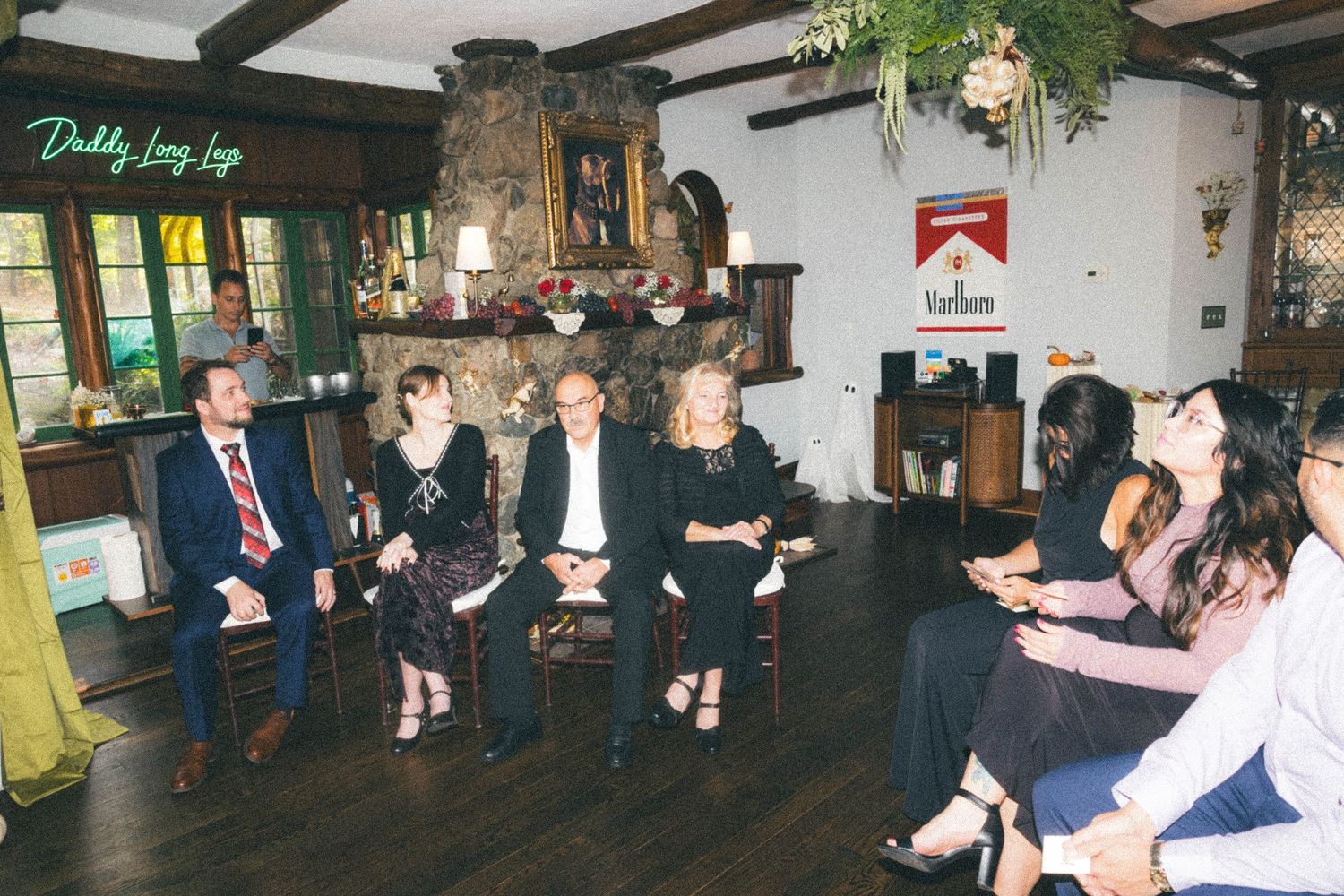 Group of wedding guests seated in rustic lodge interior with stone fireplace during reception gathering.