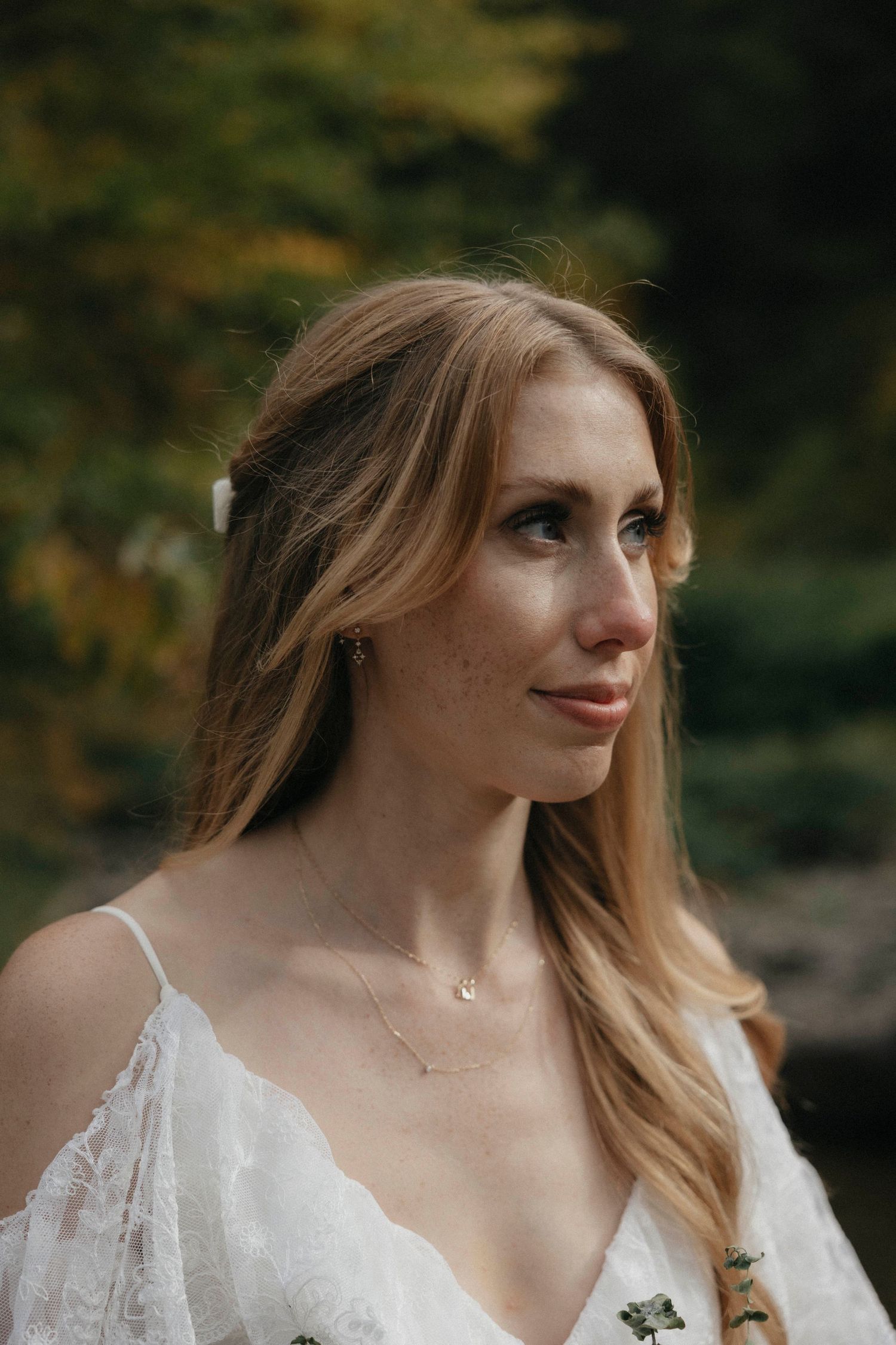Close-up portrait of blonde bride in white lace dress with delicate necklace outdoors.