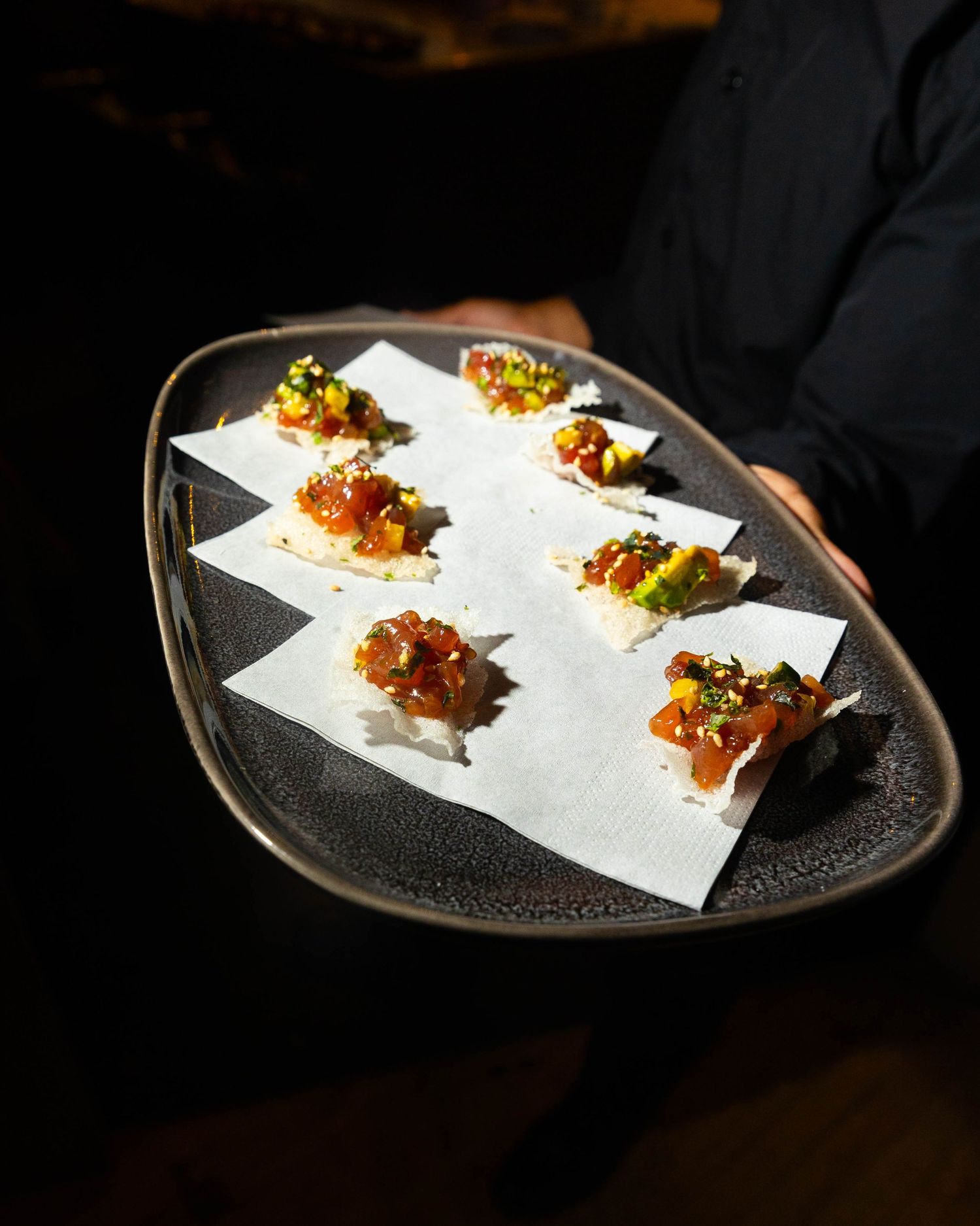 Server holding a dark ceramic tray of tuna tartare appetizers topped with avocado and garnishes on crisp cracker squares.