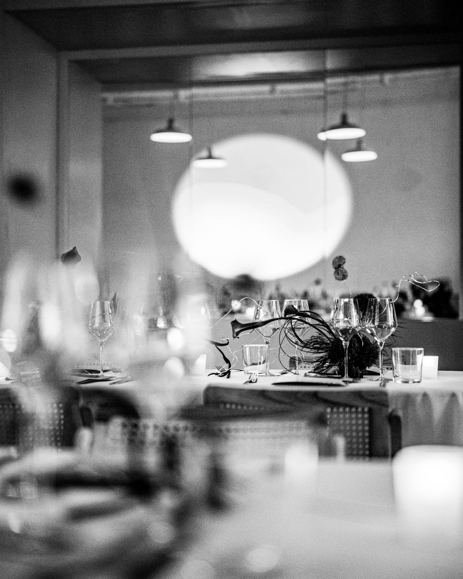 Black and white photo of an elegantly set restaurant table with wine glasses and a feathered centerpiece beneath pendant lights.