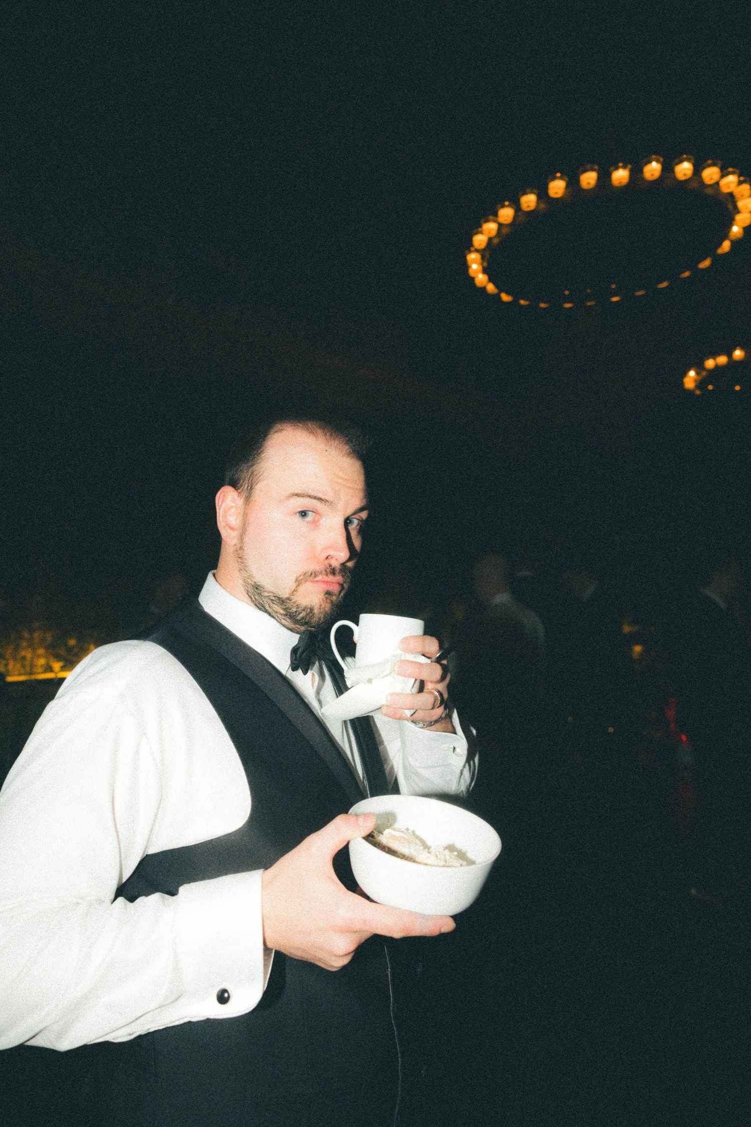 A man in a tuxedo vest holds a coffee cup and dessert bowl at an elegant evening reception.
