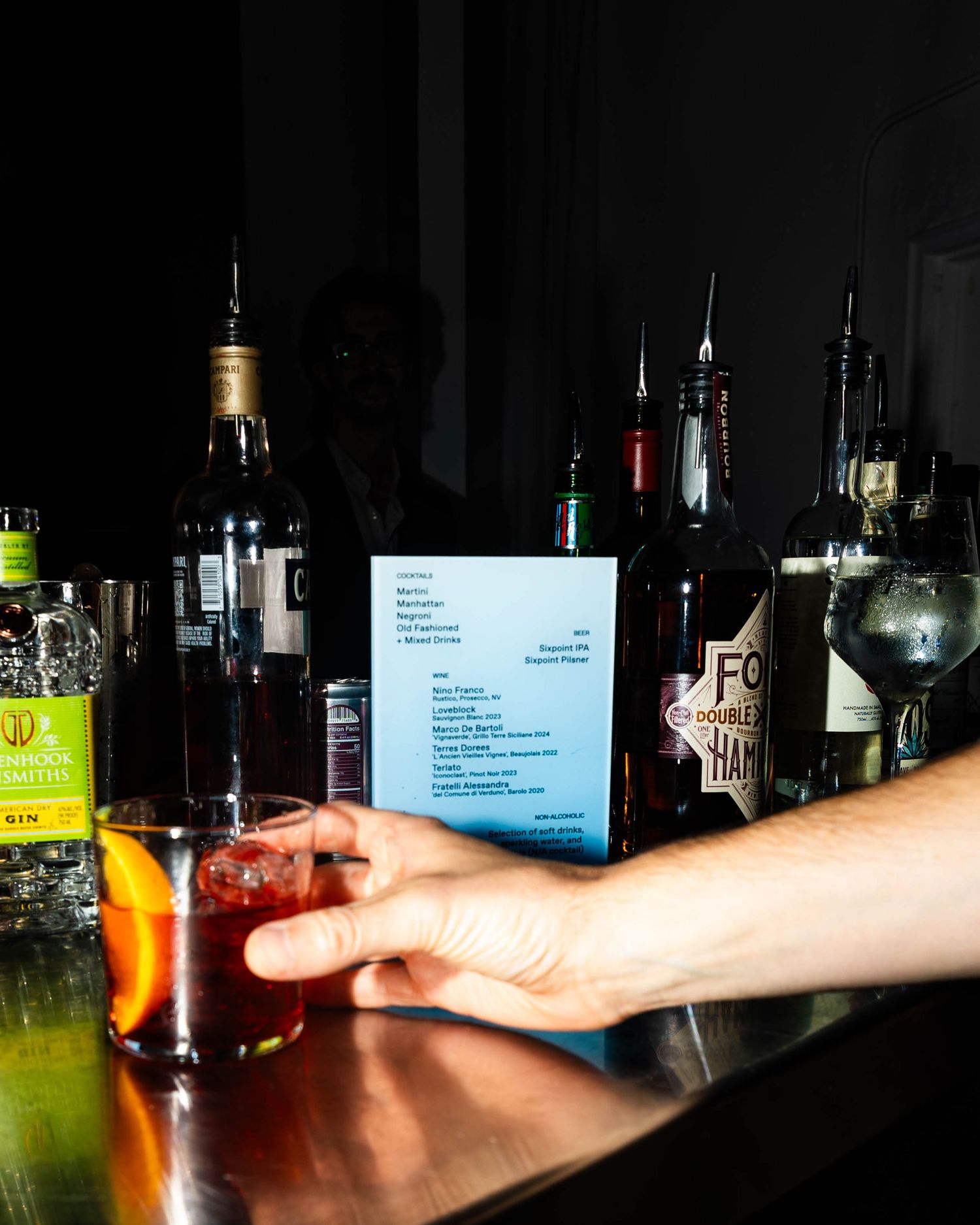 A hand reaches for a red cocktail with an orange slice on a dark bar counter, with liquor bottles and a drink menu in the background.