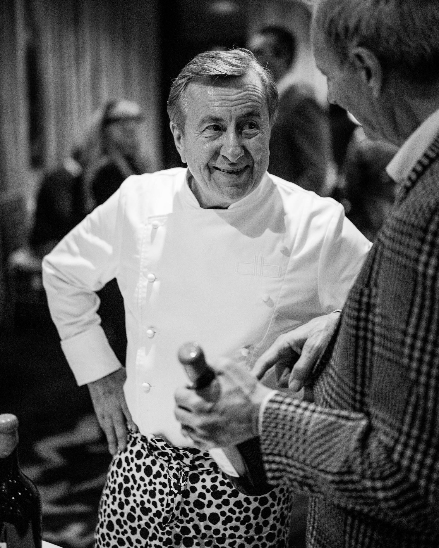 Smiling chef in a white double-breasted jacket and spotted pants chats with a man in a tweed blazer holding a wine bottle.