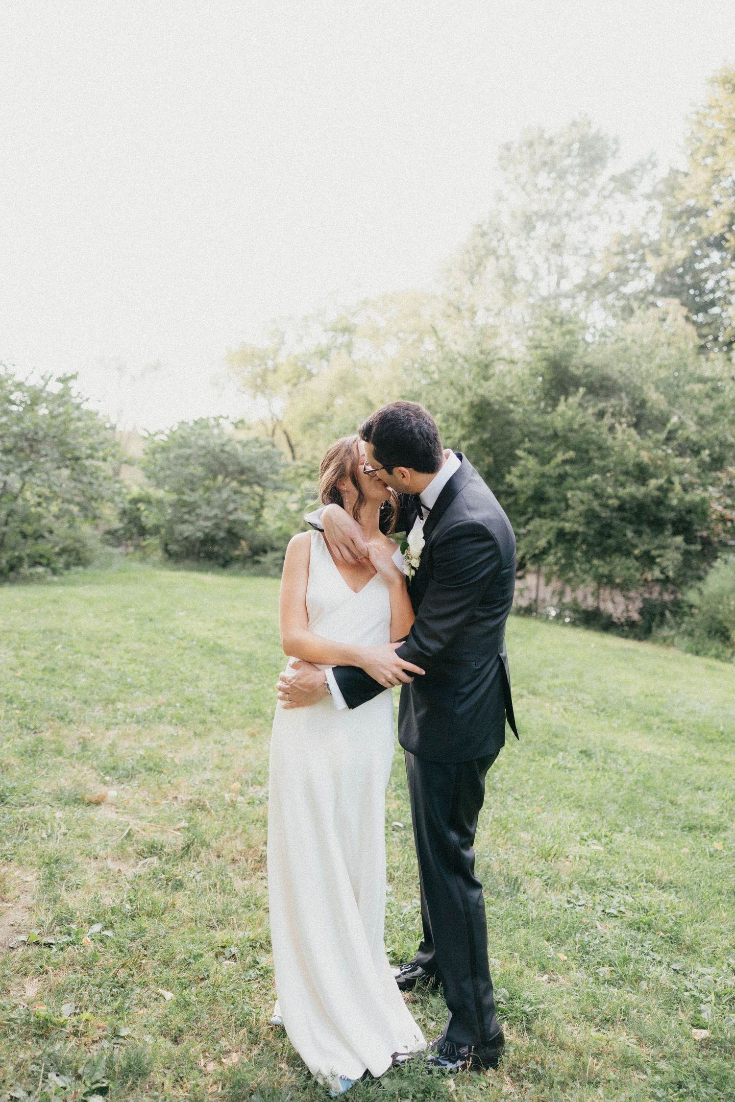 Bride and groom sharing a romantic kiss on a lush green lawn surrounded by trees on their wedding day.