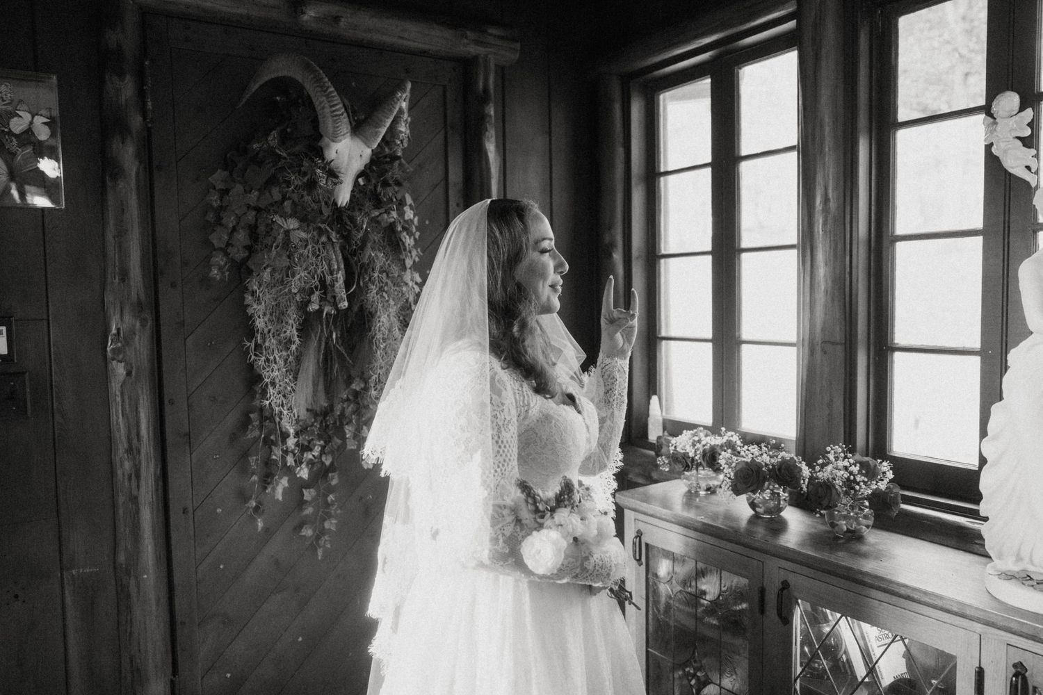 Black and white photo of bride in lace gown and veil standing by rustic wooden door.