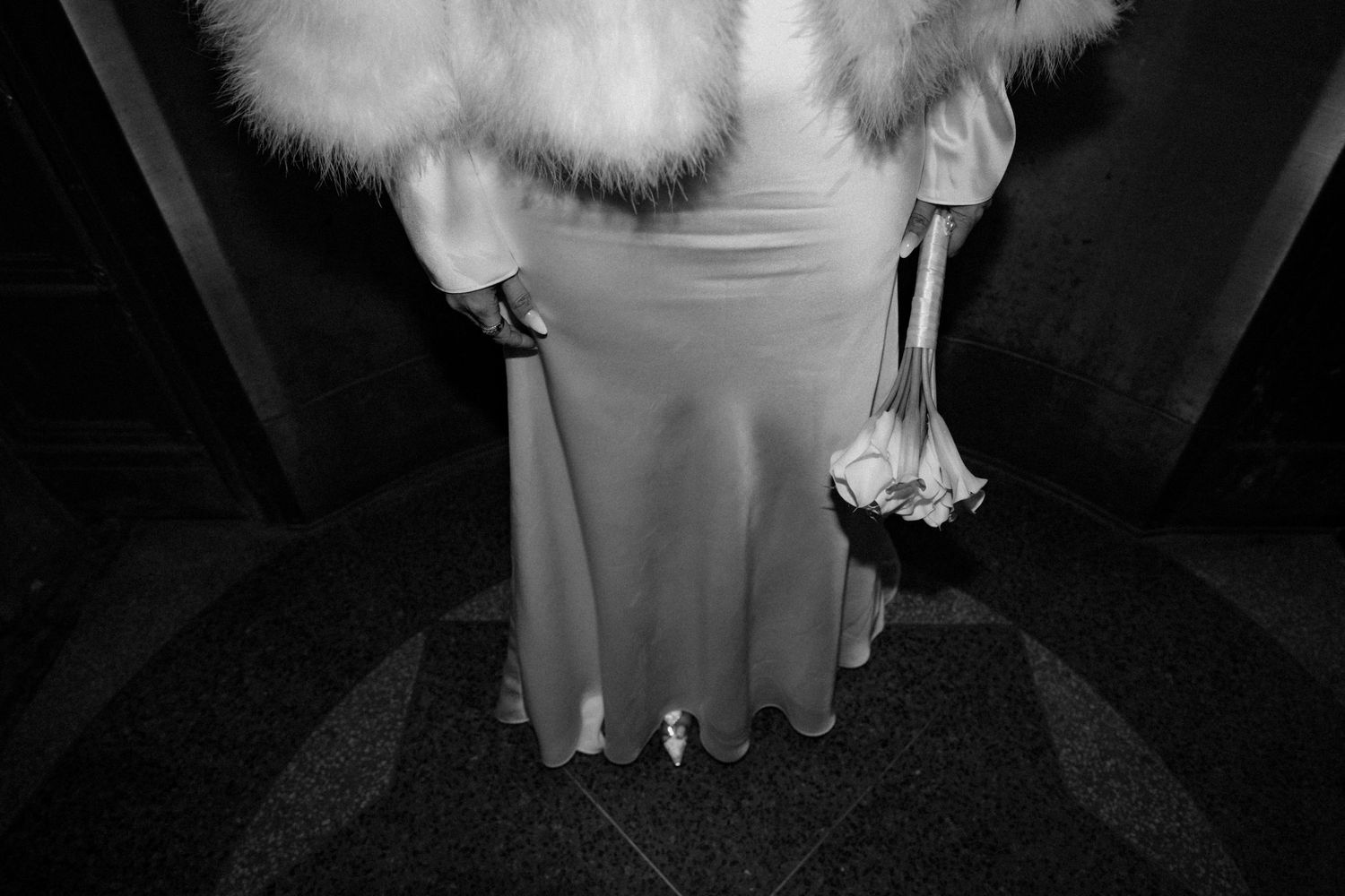 Black and white photo of bride in white gown with fur stole holding calla lily bouquet.