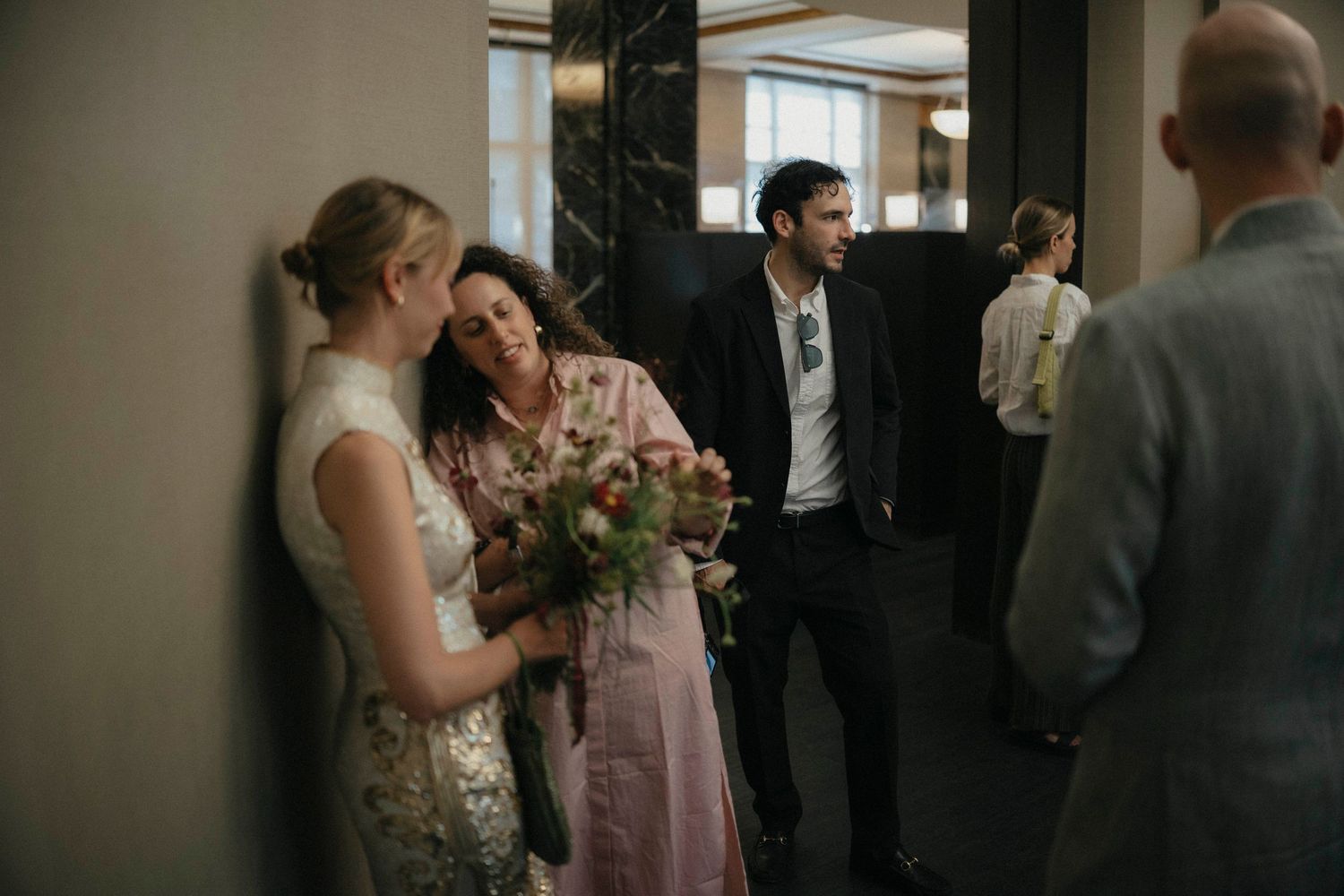 Bride in sequined dress holds bouquet while chatting with guests in a hallway.