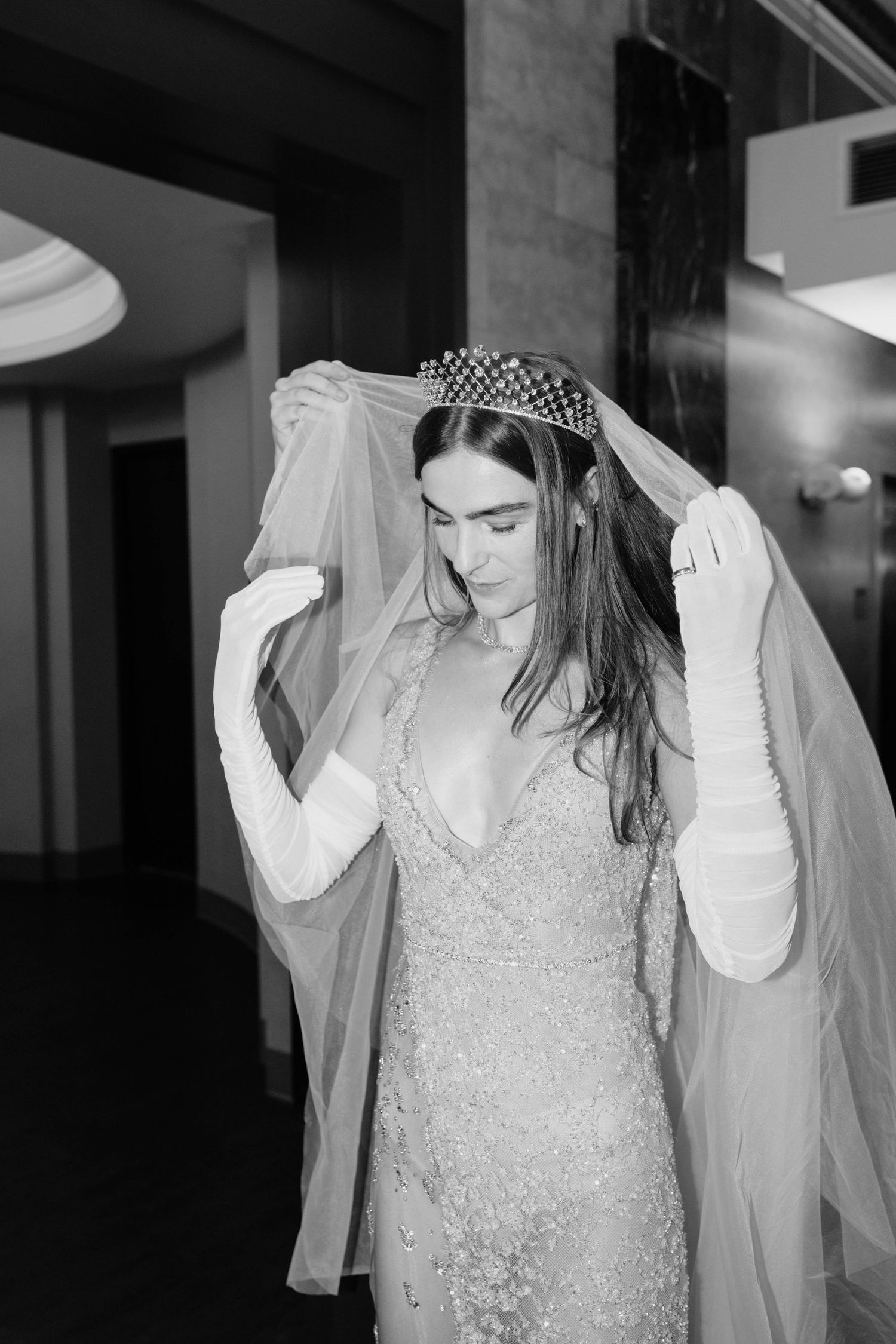 Black and white portrait of bride in beaded gown wearing pearl crown while lifting sheer veil.