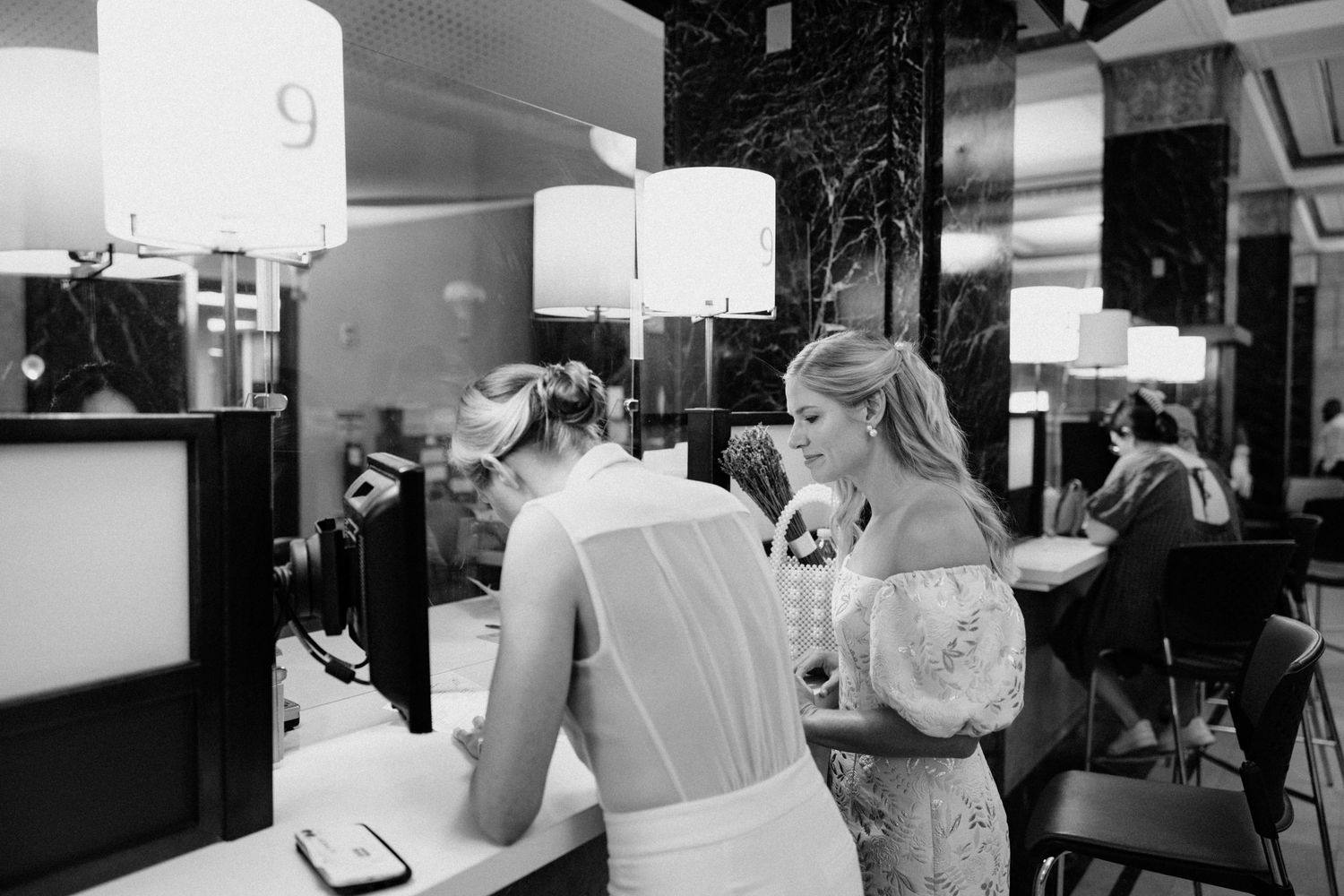 Black and white photo of two brides checking in at a hotel front desk on their wedding day.