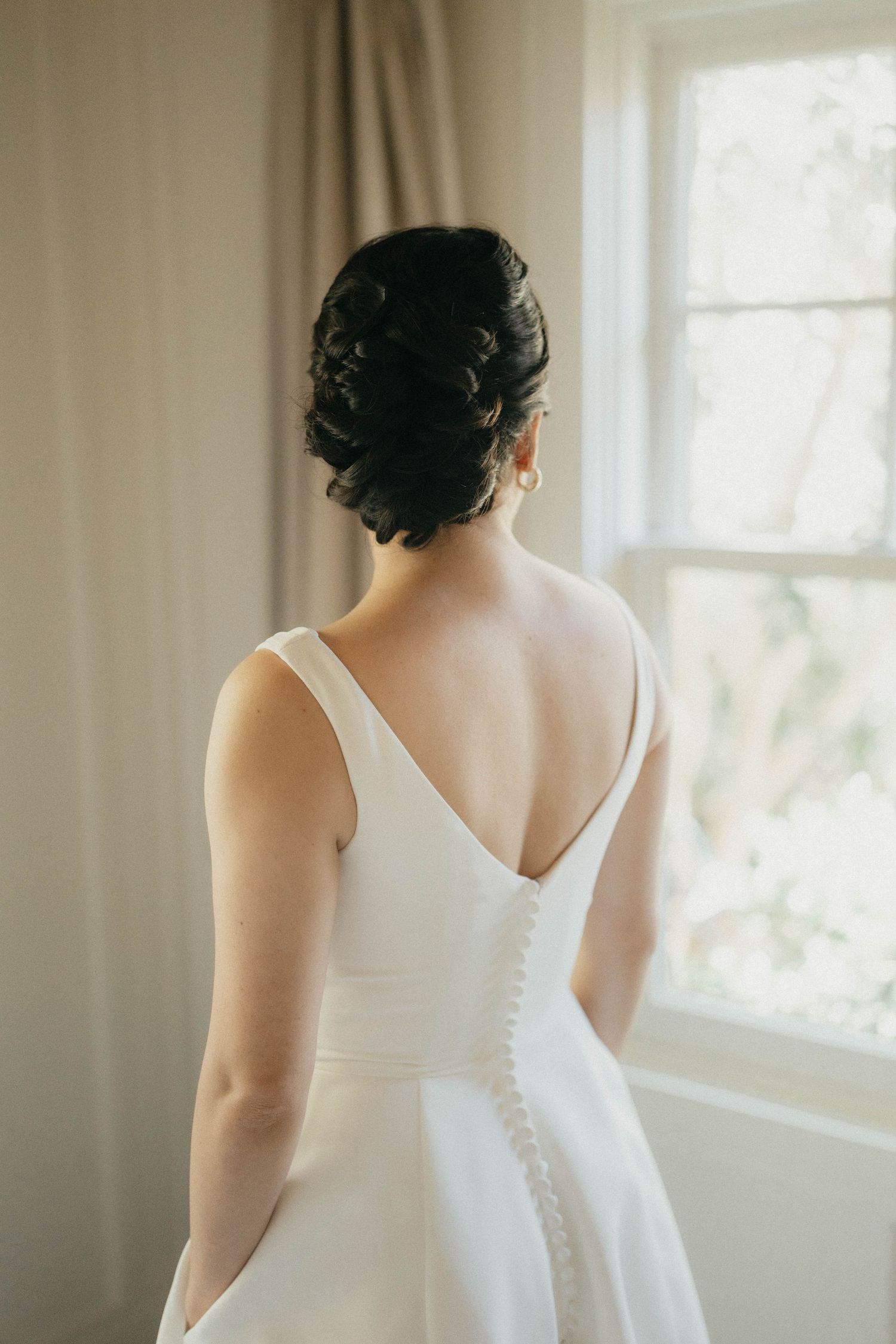 Bride with elegant updo showcases V-back wedding dress while gazing out a bright window.