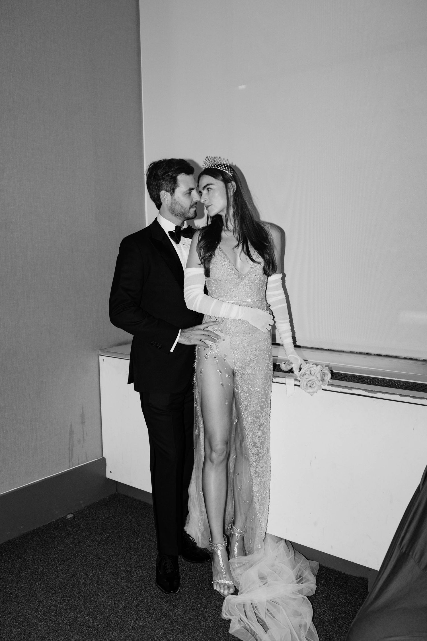 Black and white photo of newlyweds embracing, bride in sheer lace gown with crystal tiara and gloves.
