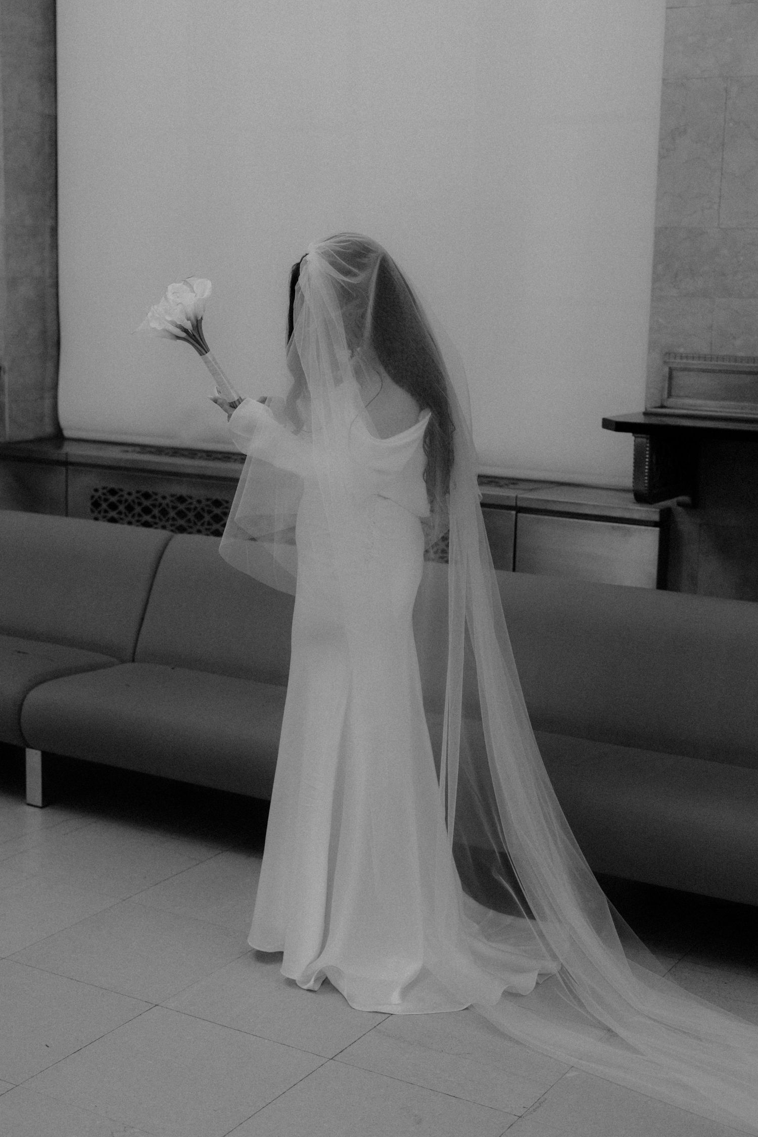 Bride in elegant white gown and long veil holds bouquet, captured in black and white.