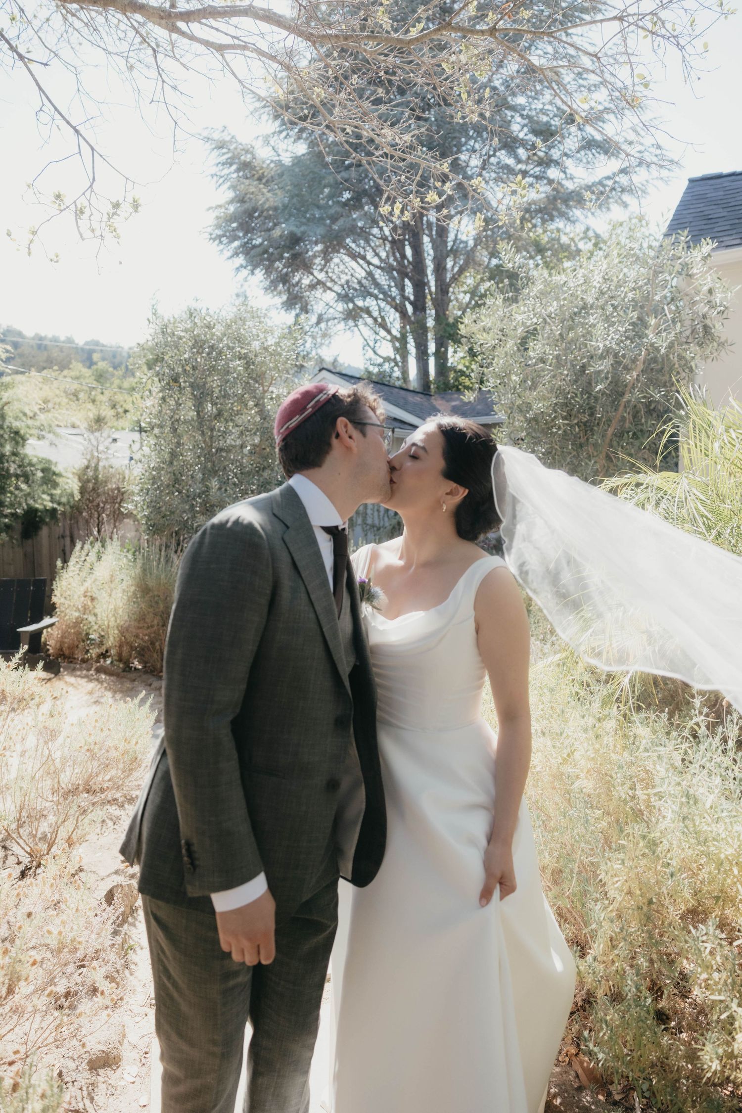 Bride and groom share a tender kiss outdoors with veil billowing in the wind.