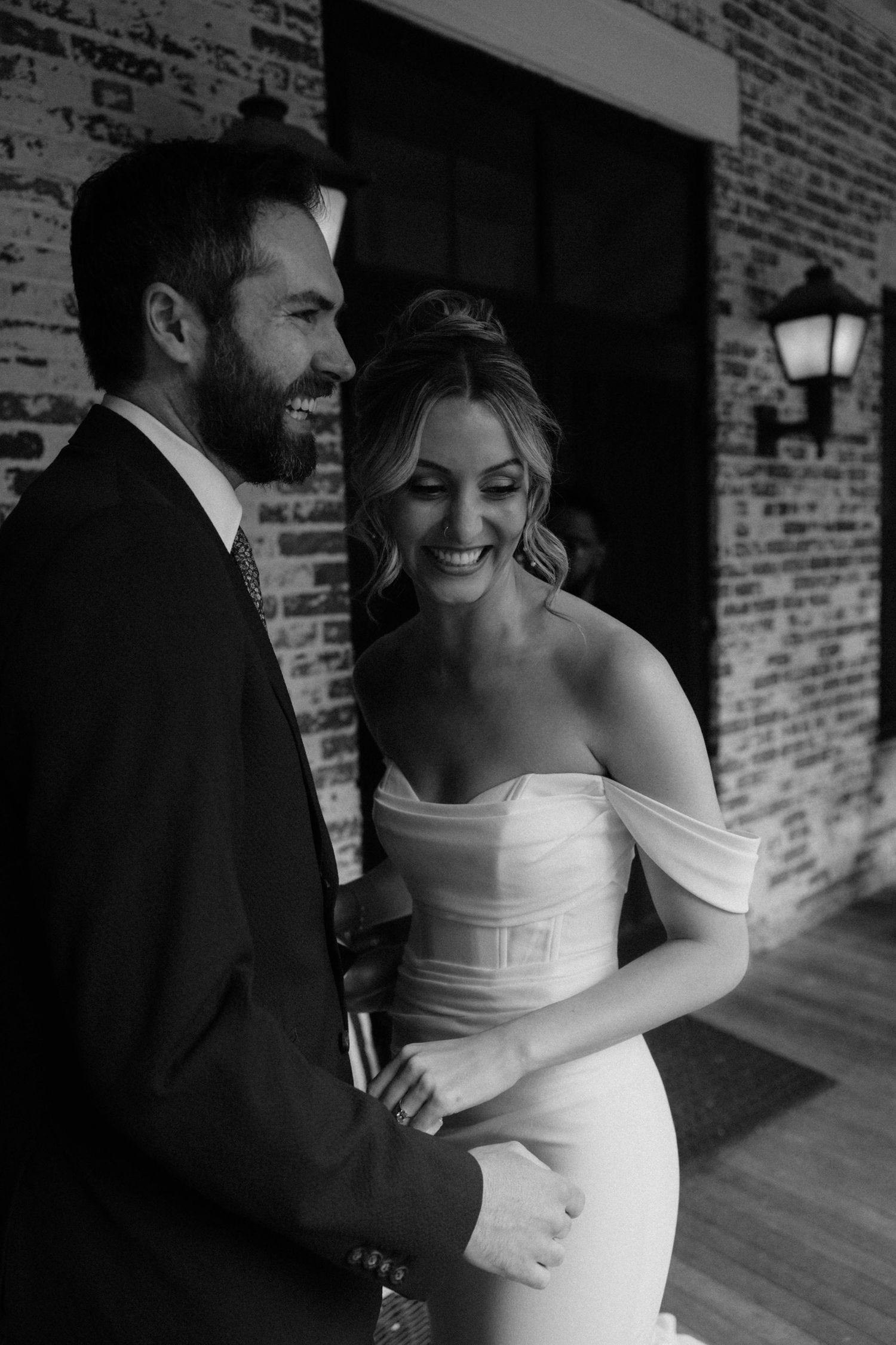 Black and white photo of a laughing bride in off-shoulder gown and bearded groom in suit at a brick wedding venue.
