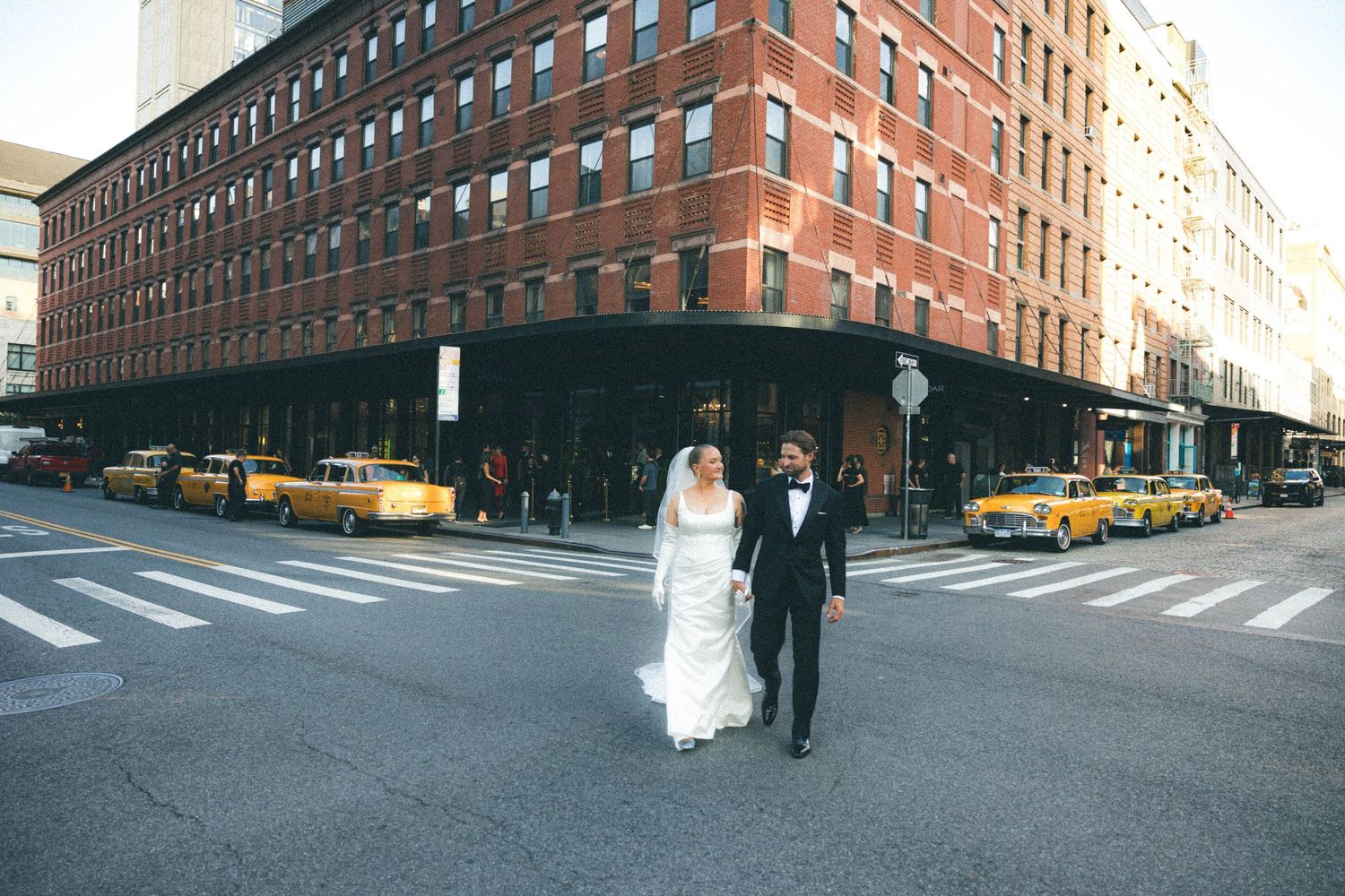 Bride and groom cross a city street in New York with yellow taxis and brick buildings behind them.