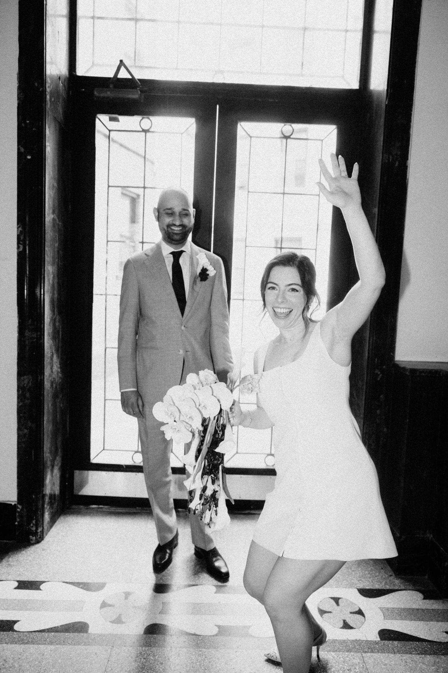 Joyful bride in white mini dress waves bouquet while groom smiles in black and white photo.