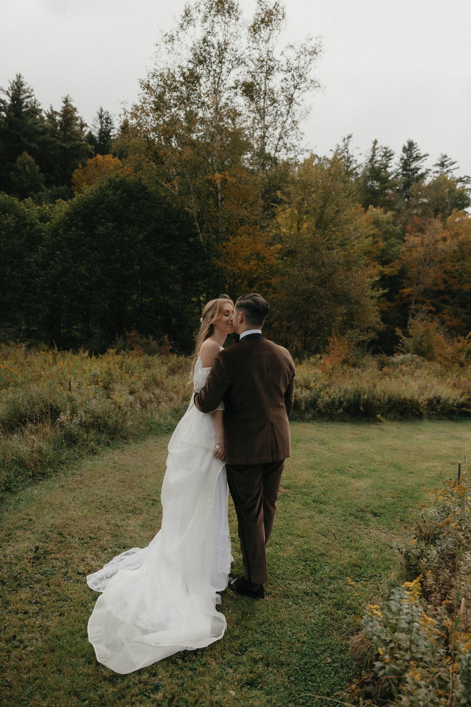 Bride and groom sharing a kiss on green lawn with vibrant autumn foliage backdrop.