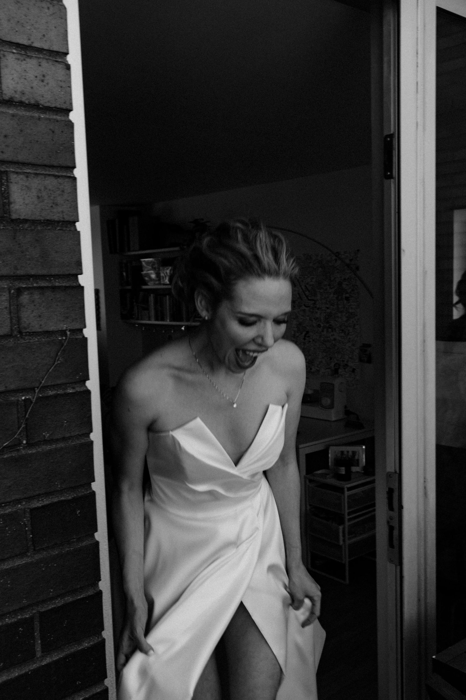 Bride in strapless white satin gown laughing joyfully in doorway during wedding preparations, black and white photo.