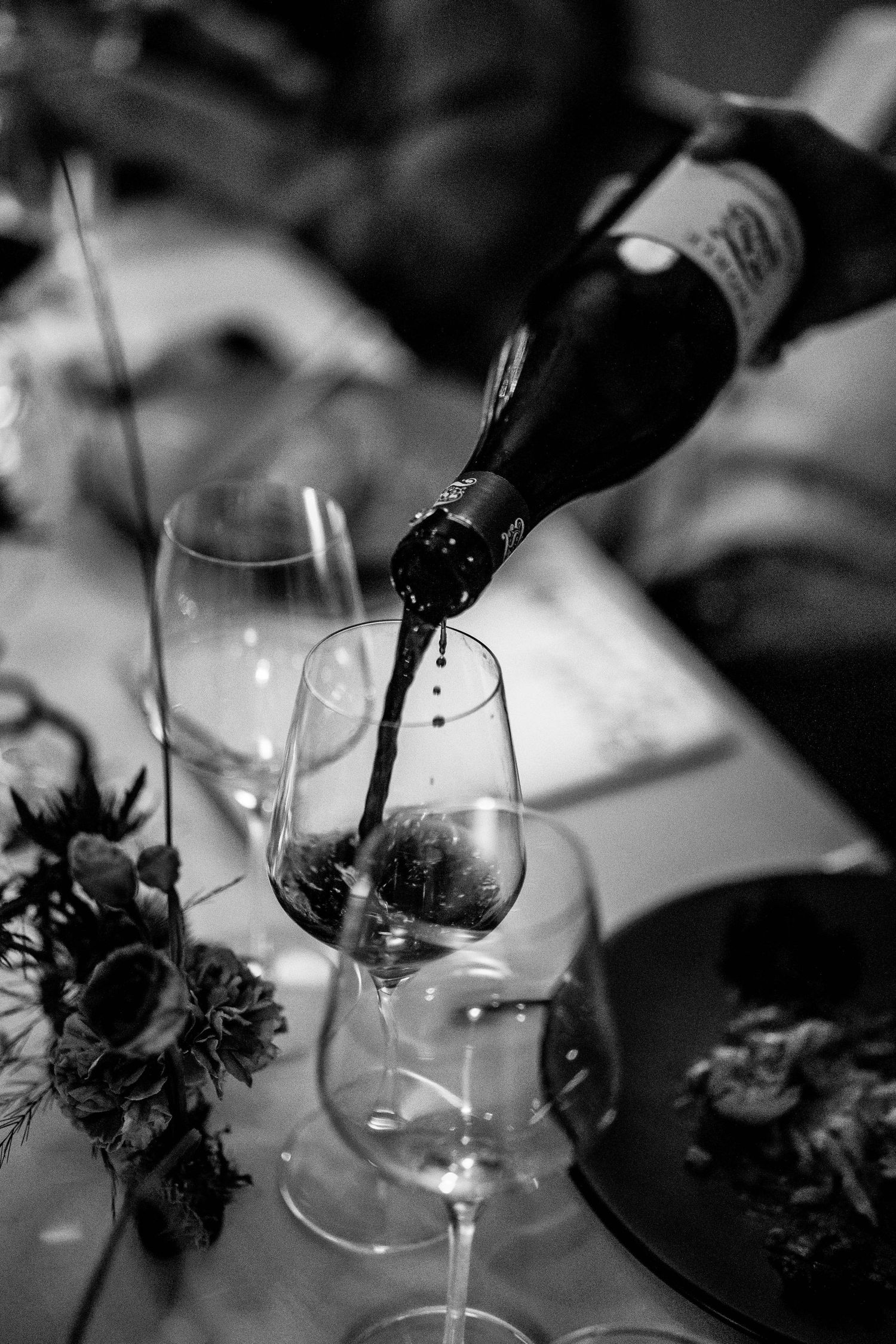 Black and white photo of red wine being poured from a bottle into a glass at a elegantly set dinner table.